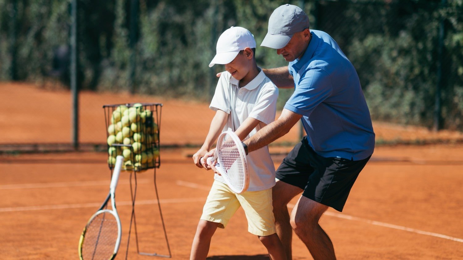 An adult and a child practicing tennis on a clay court with tennis rackets, with a basket of tennis balls nearby.