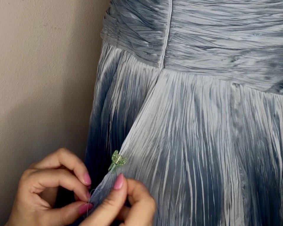 Close-up of a person sewing a small decorative dragonfly onto a silver and blue pleated dress.
