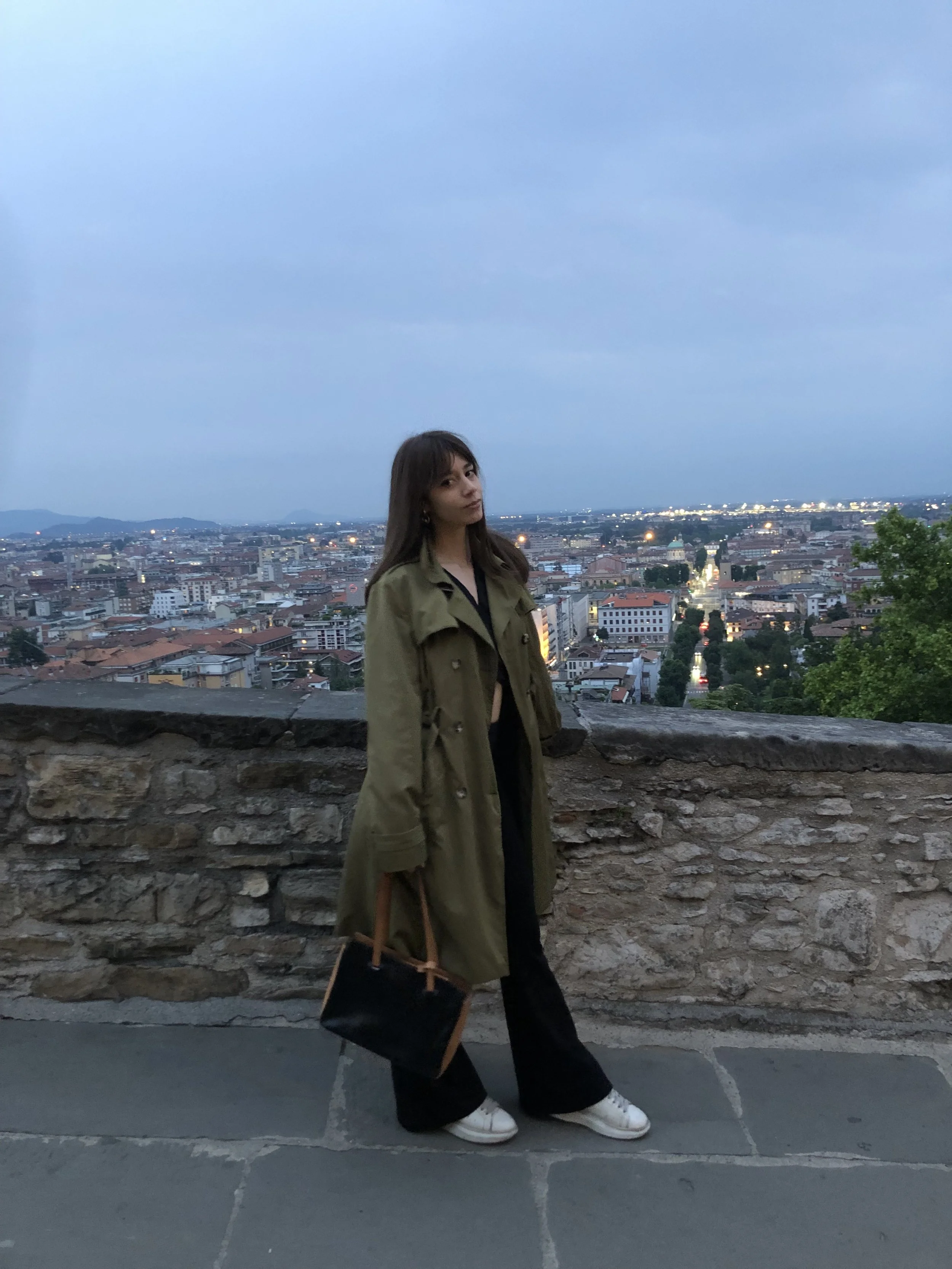 A woman standing in front of a cityscape during dusk, wearing a green trench coat, black pants, white sneakers, and carrying a black handbag.