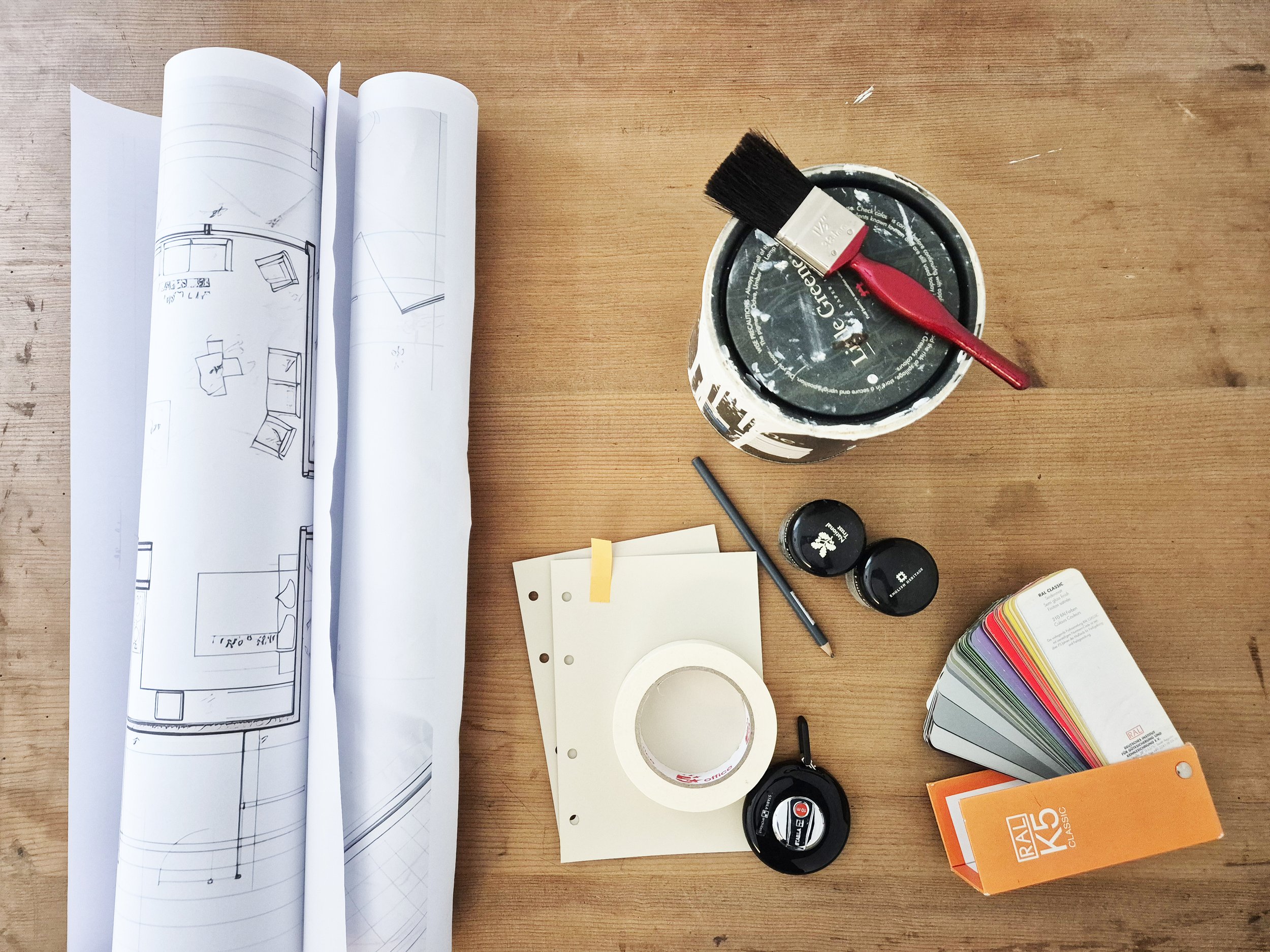 A wooden desk with blueprints, a paint can with a paintbrush, color sample cards, and various small tools and paint containers.
