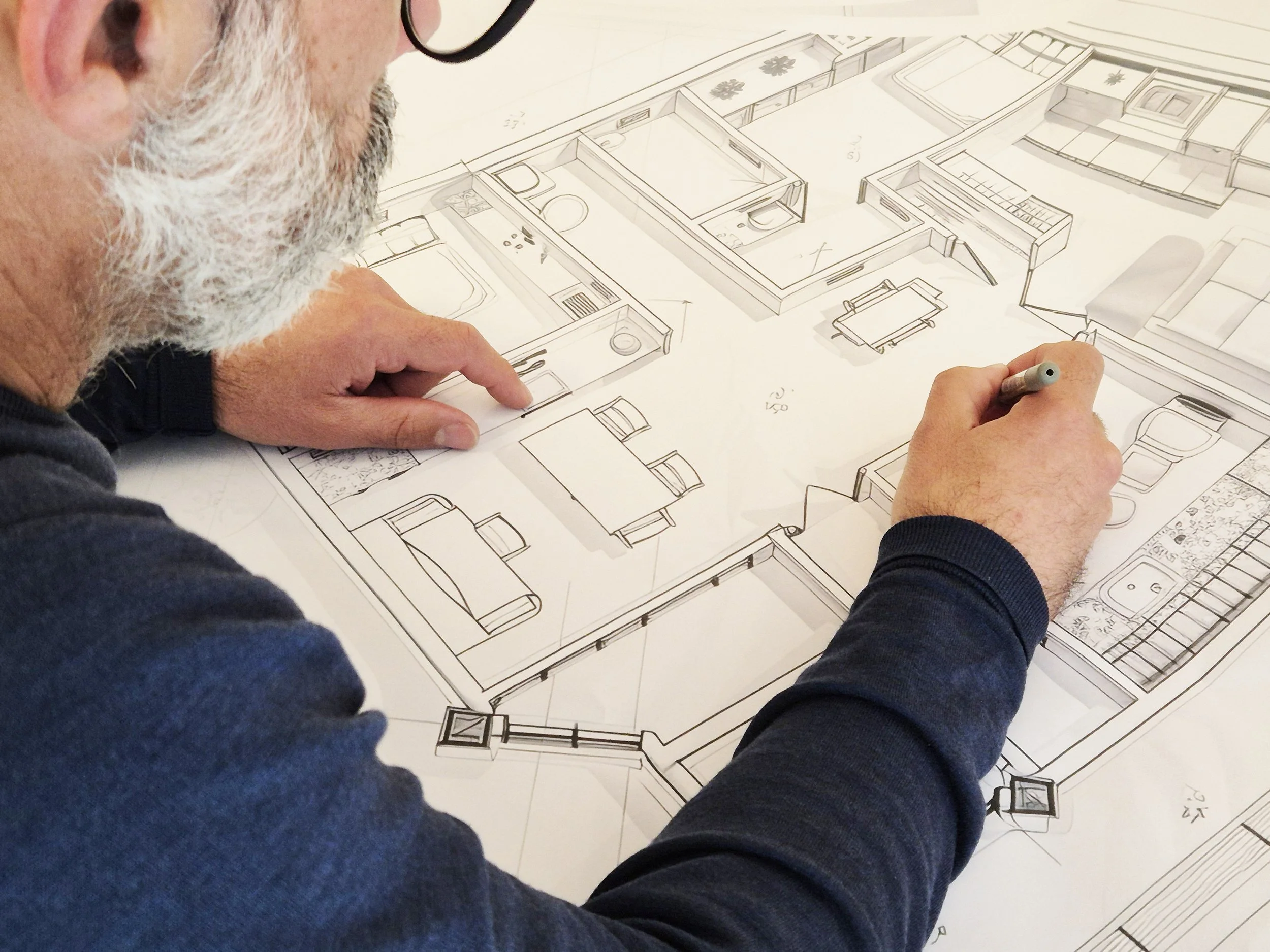 A person with a gray beard, glasses, and wearing a dark long sleeve shirt is drawing or examining a detailed architectural floor plan or blueprint of a building layout.