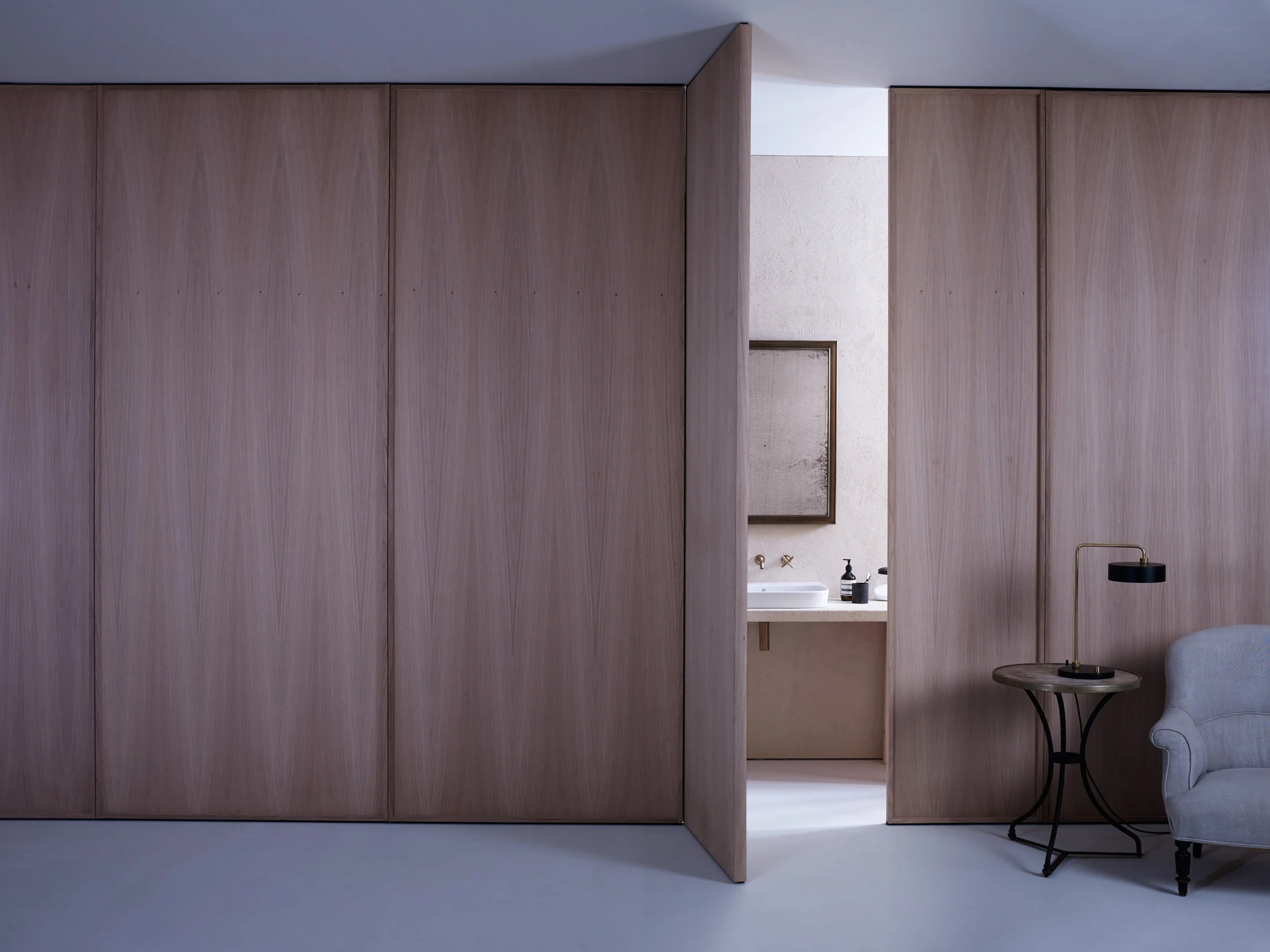 View of an interior room with a partially open wooden sliding door revealing a bathroom sink and mirror, next to a sitting area with a gray armchair, black side table, and black and gold desk lamp.