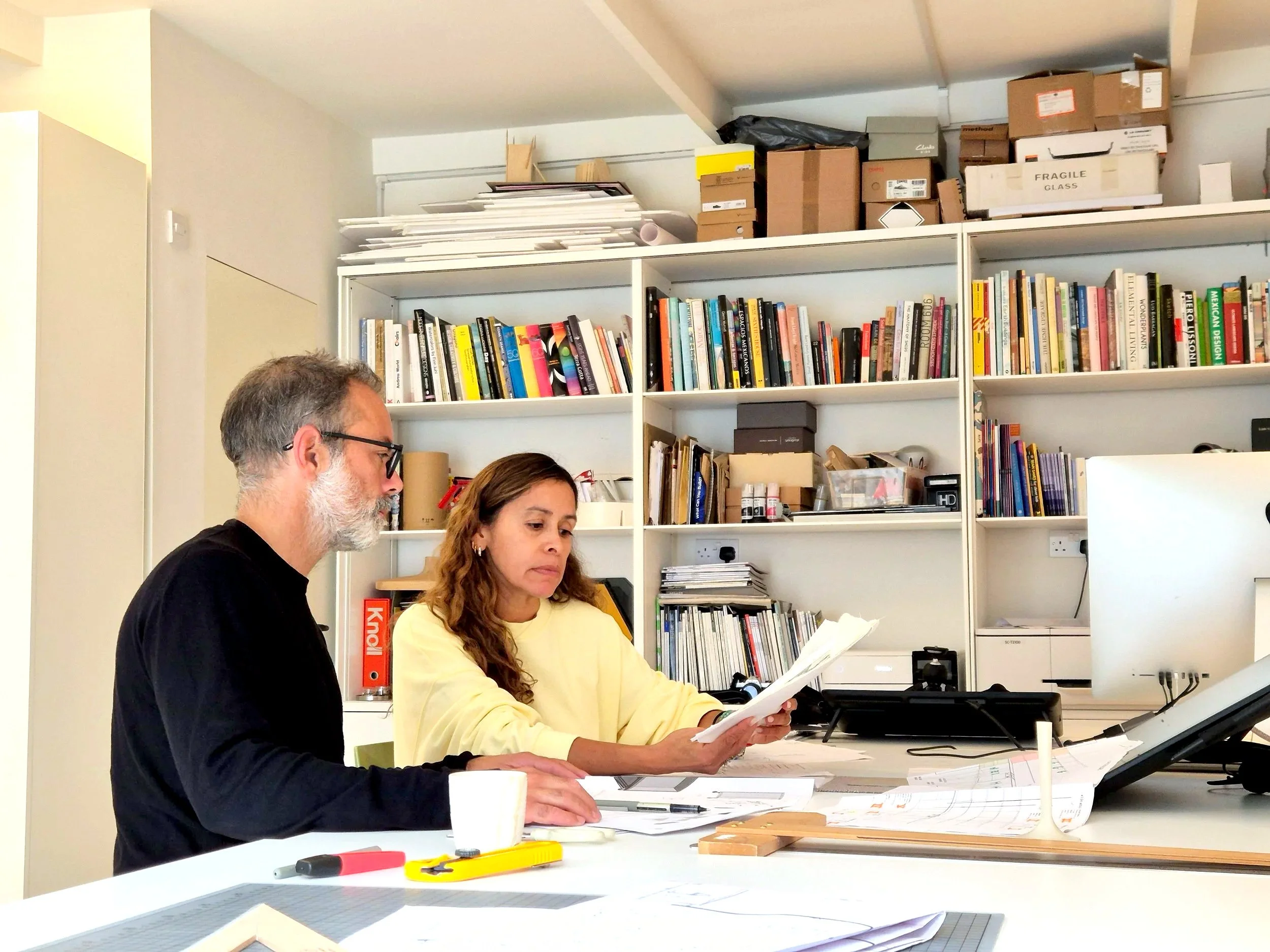 Two people, a man with glasses and a woman, working at a desk with paperwork and drawing tools, in an office filled with shelves of books, papers, and boxes.