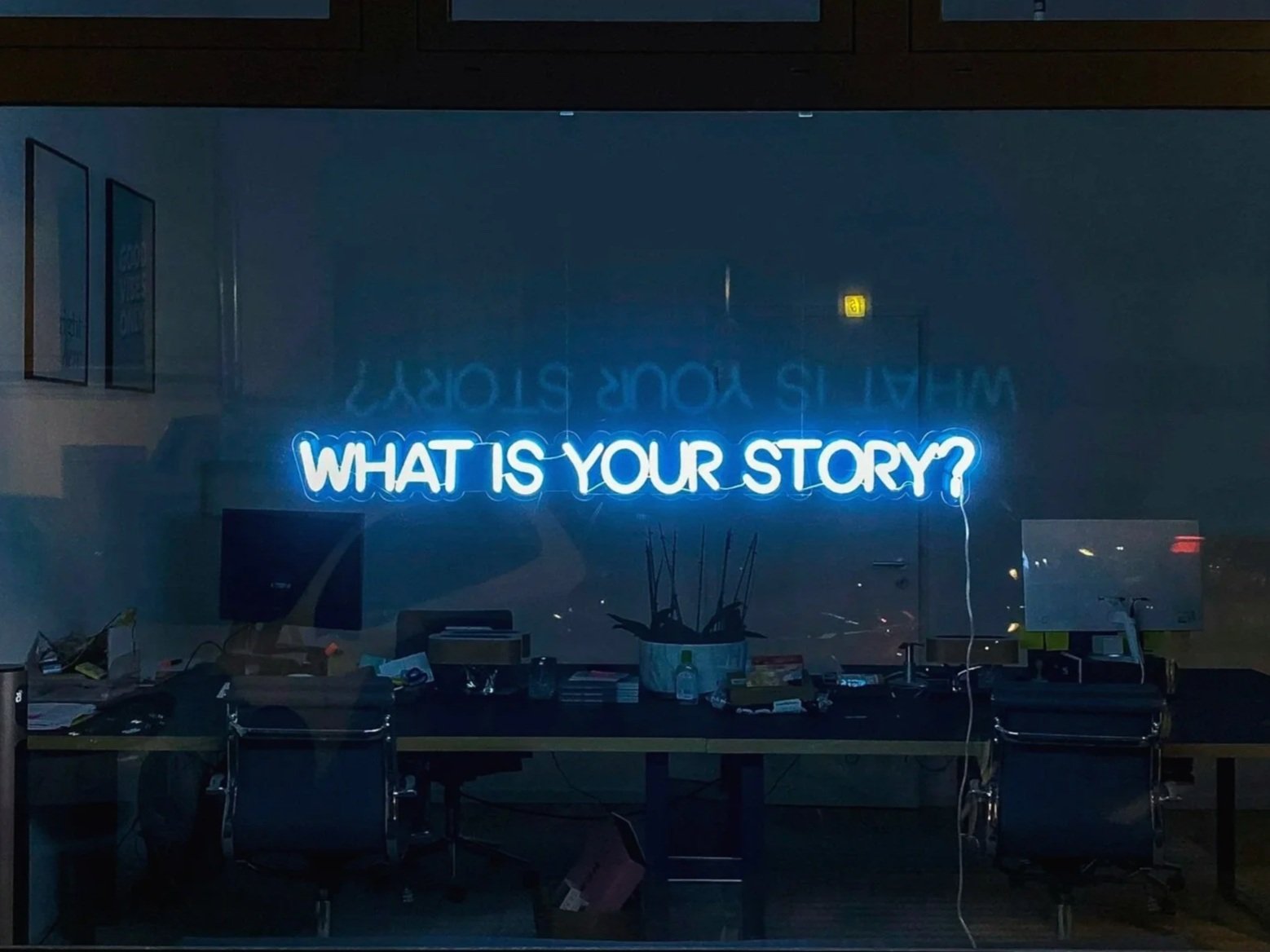 A neon sign with the text 'WHAT IS YOUR STORY?' illuminated in blue inside a dark office space.