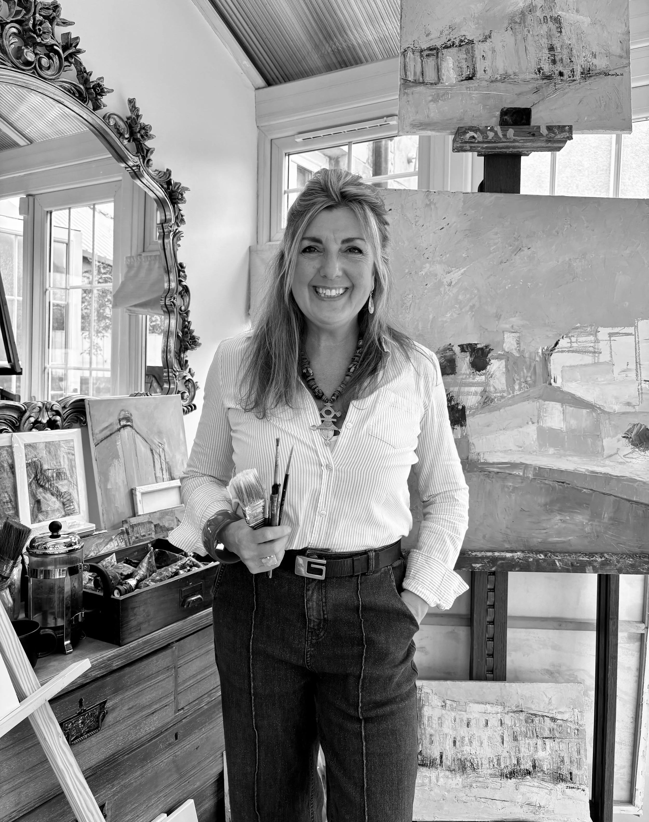 A smiling woman stands in an art studio holding paintbrushes, surrounded by paintings and art supplies, with a large mirror and window in the background.