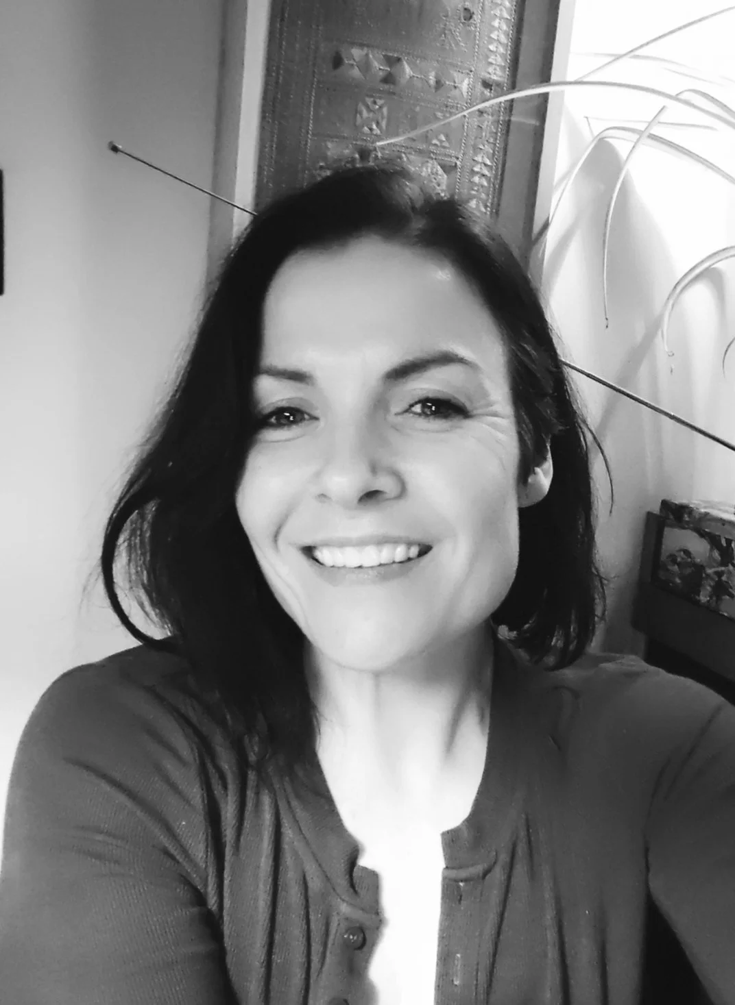 A woman with shoulder-length dark hair smiling and taking a selfie in a room, with a patterned wooden wall hanging and a plant in the background.