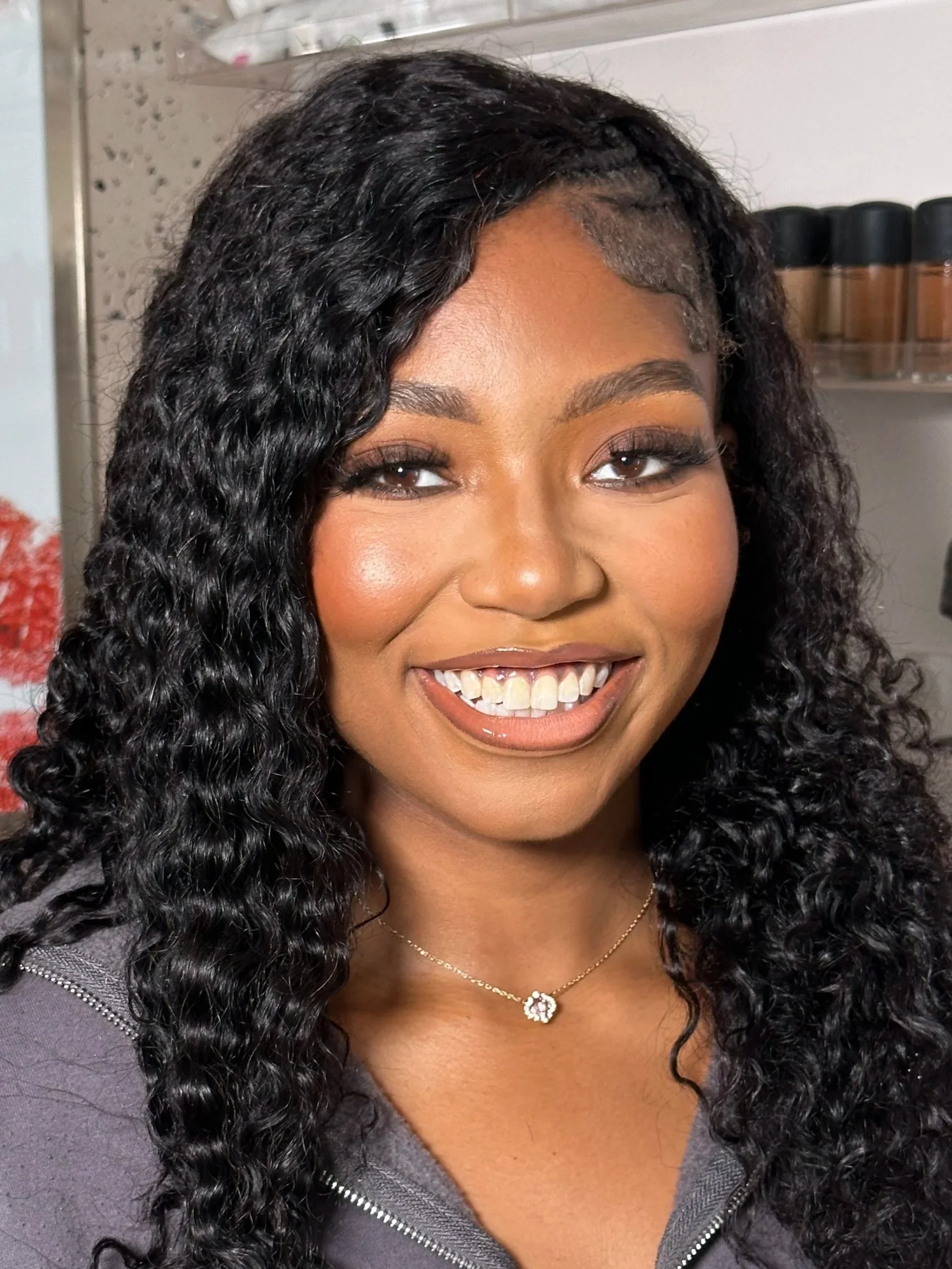 A smiling woman with long, curly black hair, wearing makeup, a delicate necklace, and a gray top.