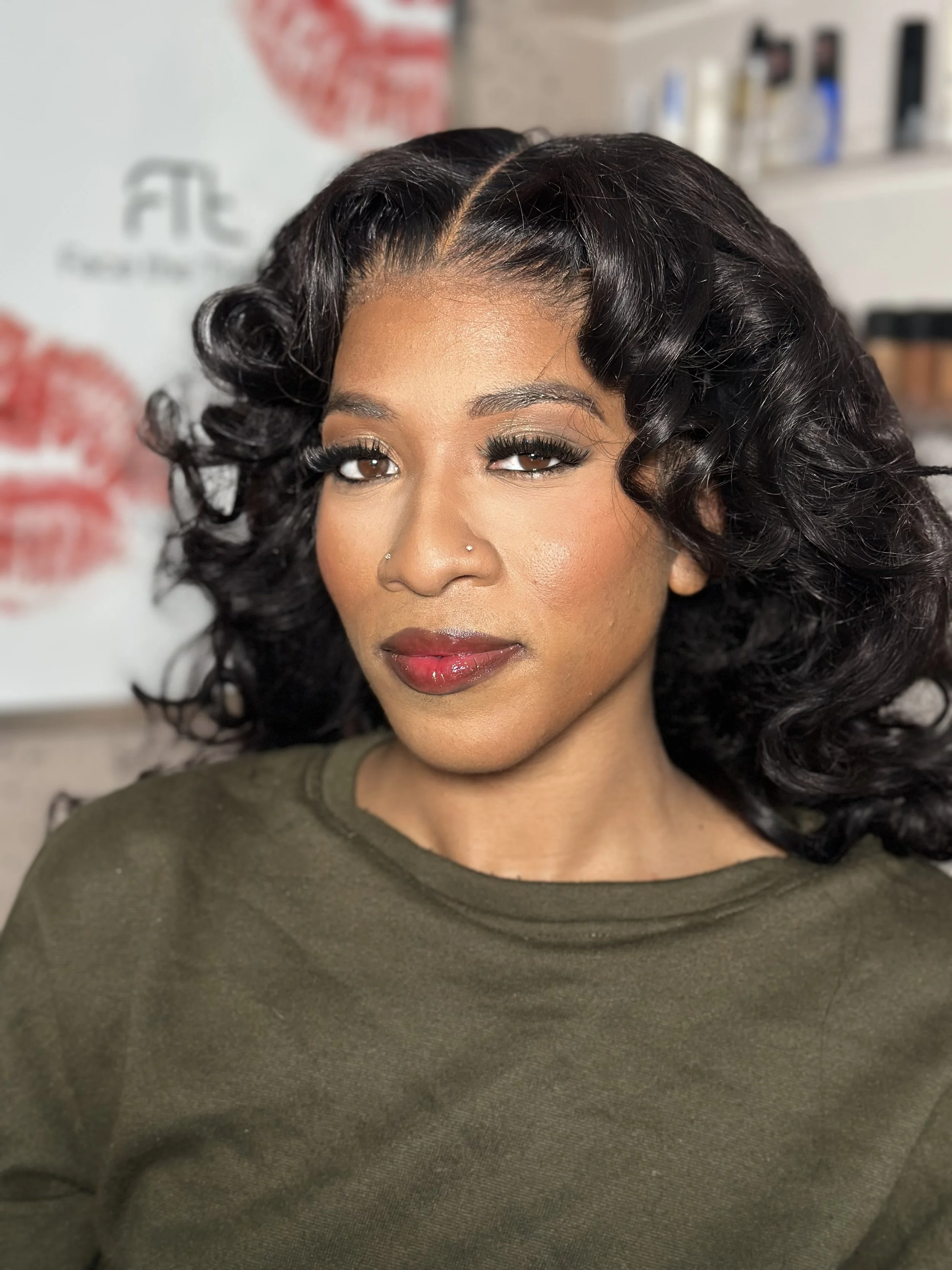 Woman with curly black hair, makeup, and a nose piercing, wearing an olive green top, sitting in a salon with a blurred background.