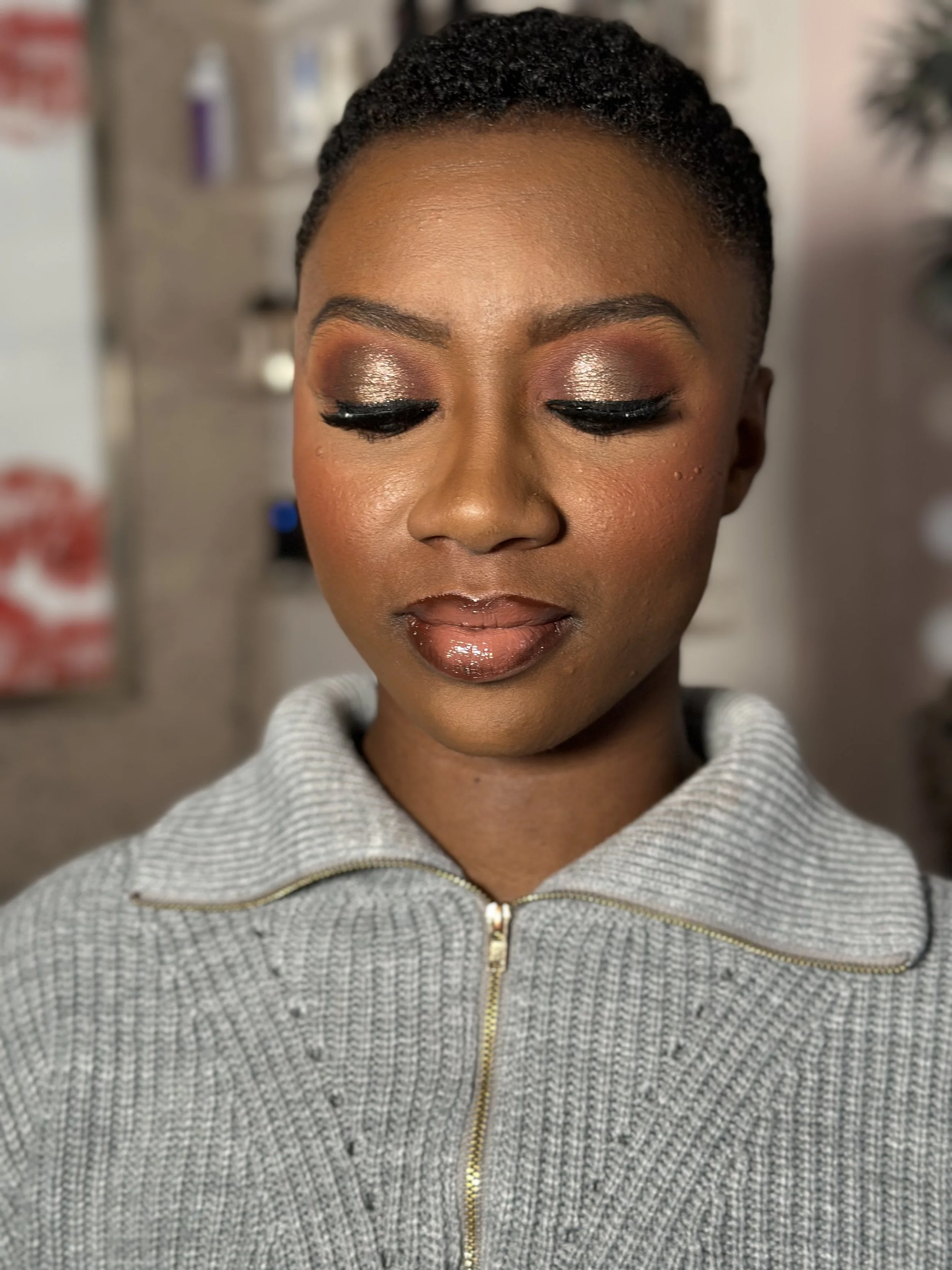 Close-up of a woman with short, natural hair wearing makeup with gold eyeshadow, winged eyeliner, and glossy lips, dressed in a gray zip-up sweater.