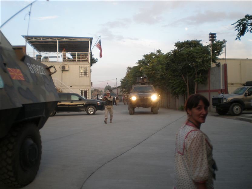 Sarah Hearn OBE is turning to smile in front of armored vehicles in a NATO compound in Afghanistan in 2010. Soldiers and Macedonia and USA flags are also visible.
