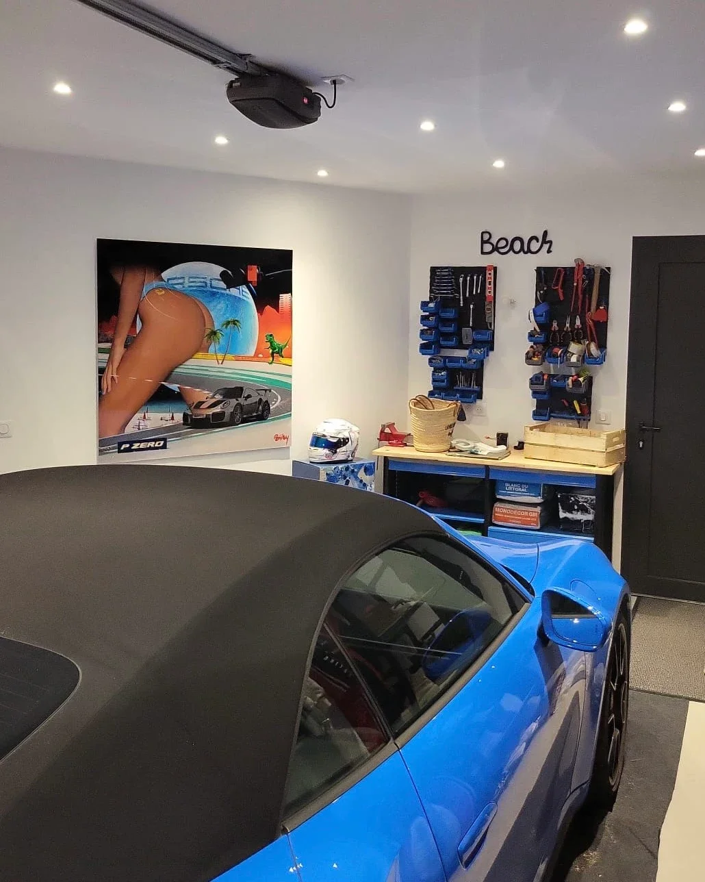 Un garage avec une voiture bleue, un mur décoré d'une affiche d'une femme en maillot de bain, un panneau avec des outils, et un panneau nommé 'Beach'.