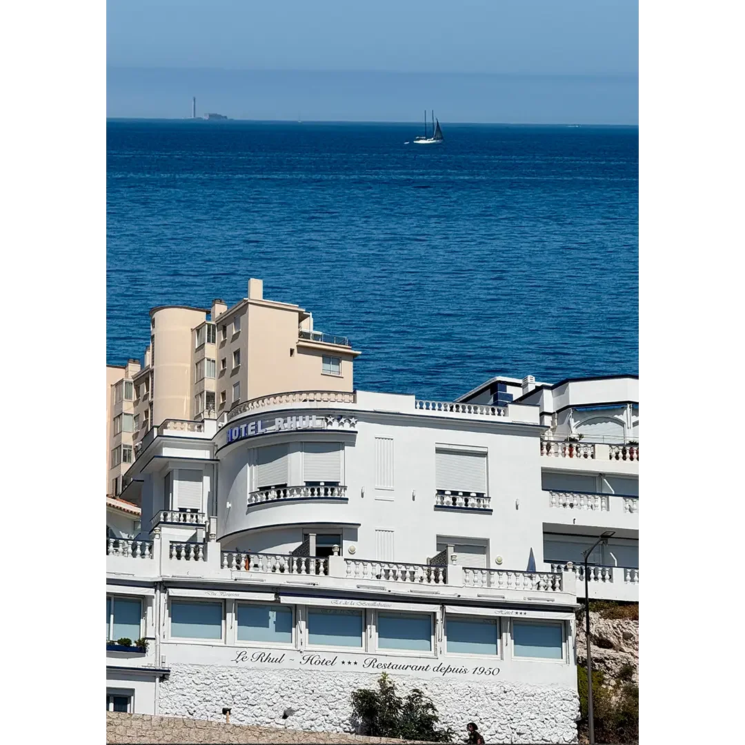 Vue d'un bâtiment blanc, nommé Hôtel Le Ruhl, avec vue sur la mer où naviguent un voilier et un autre bateau.