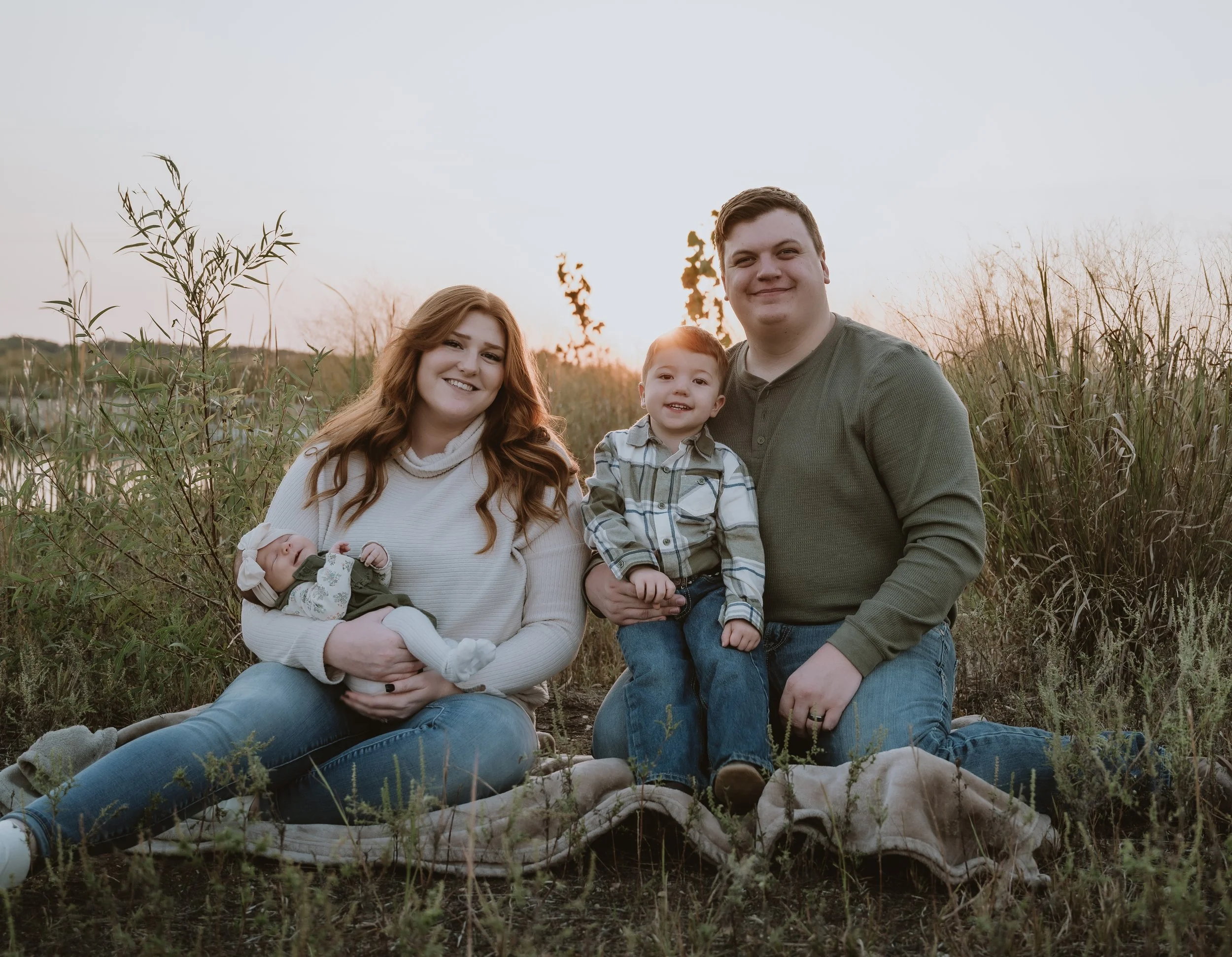 A family of four sitting outdoors in a grassy field at sunset, smiling at the camera. The mother is holding a newborn, and a toddler is sitting between the father and mother.
