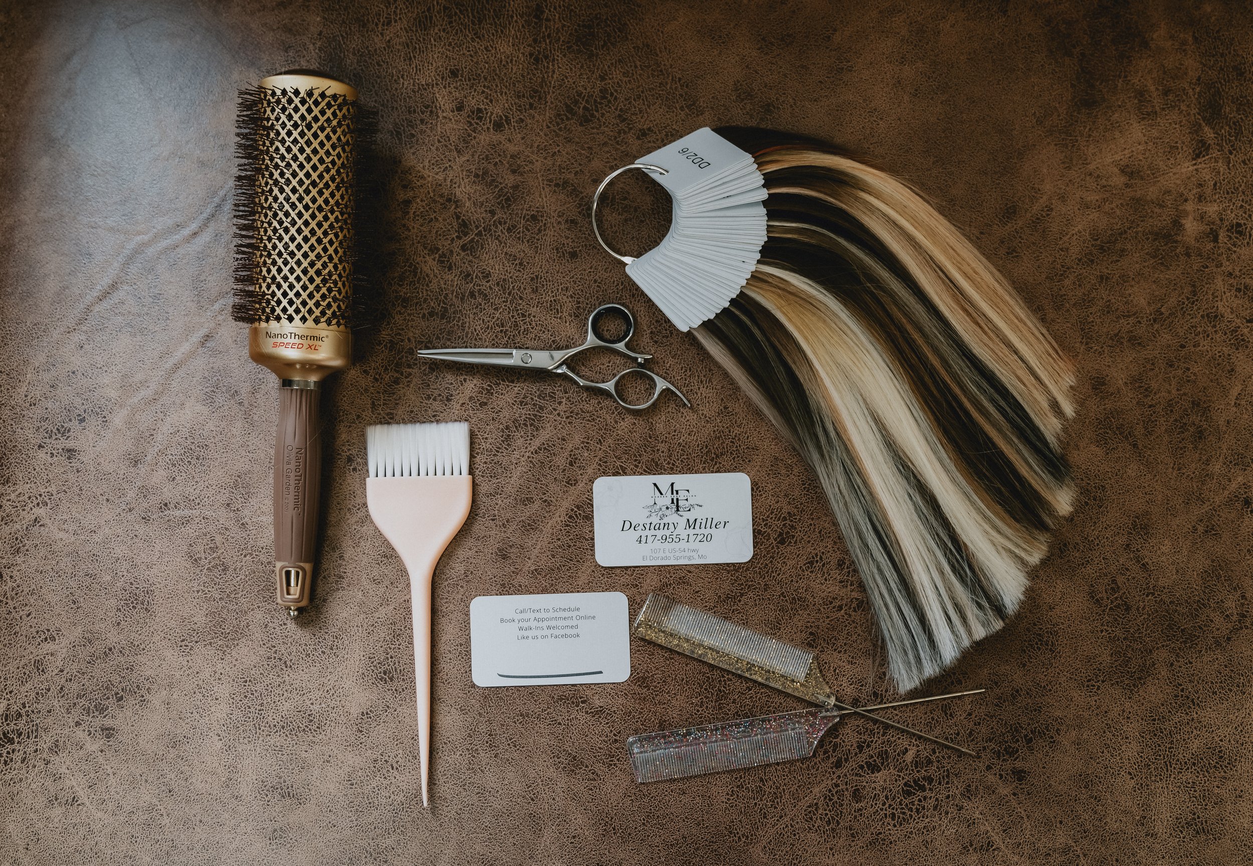 Hair coloring tools and samples including a hairbrush, scissors, a hair color fan with hair sample swatches, a small hairbrush, business cards, a glittery hair clip, and a decorative hairpin on a brown textured surface.