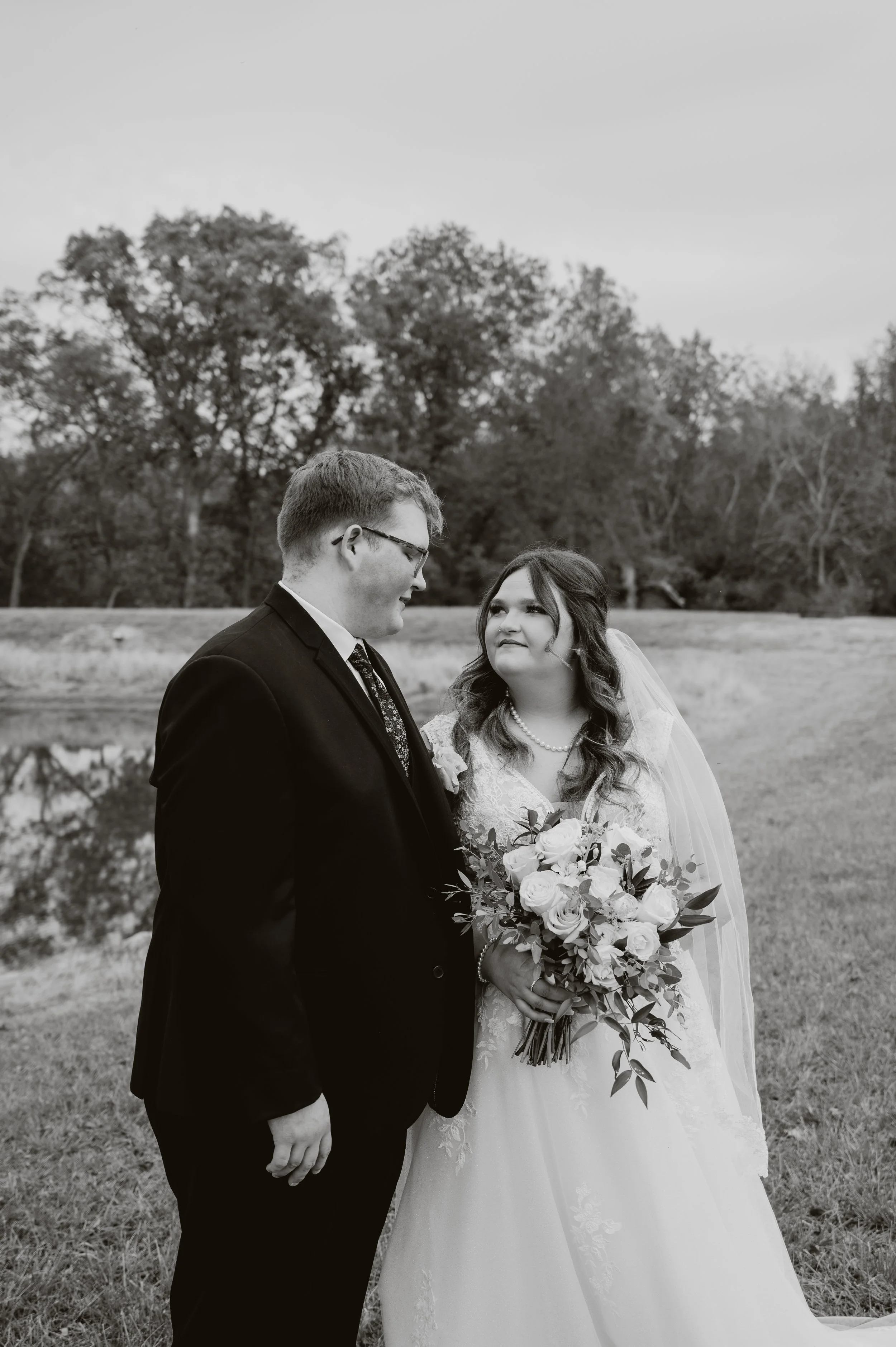 Black and white portrait of a bride and groom by a quiet pond, sharing an intimate moment.
