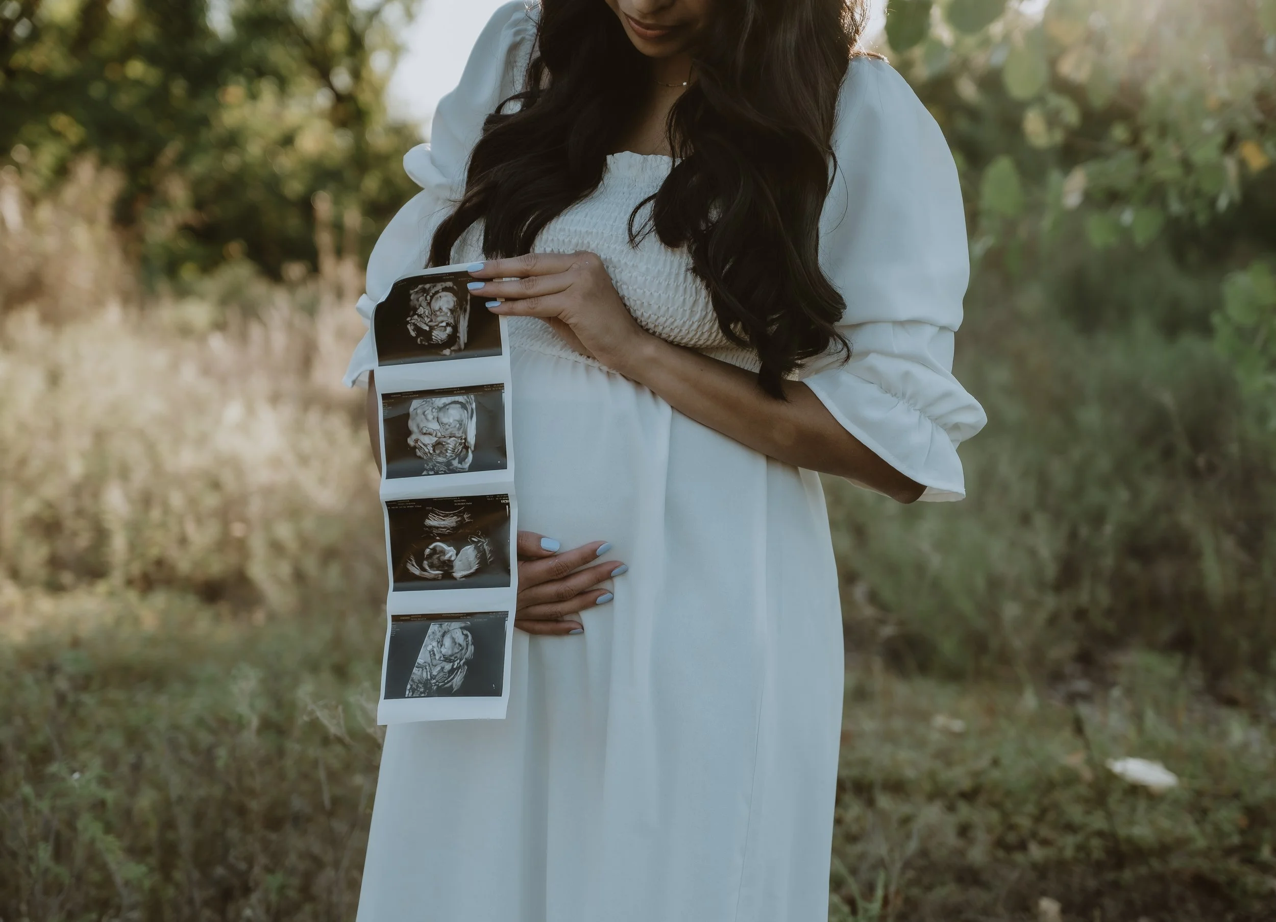 A pregnant woman in a white dress holding her belly with one hand and a string of ultrasound images with the other, outdoors with green trees and sunlight in the background.