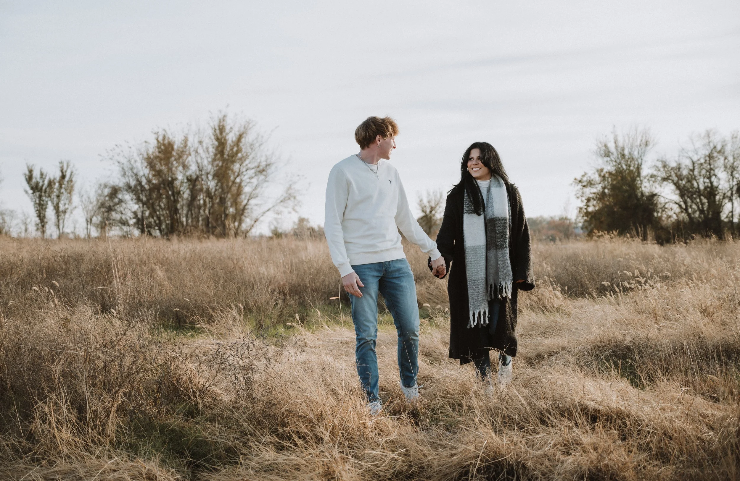 Lifestyle engagement portrait of a couple walking hand in hand through tall grass, surrounded by open land and a soft, overcast sky.