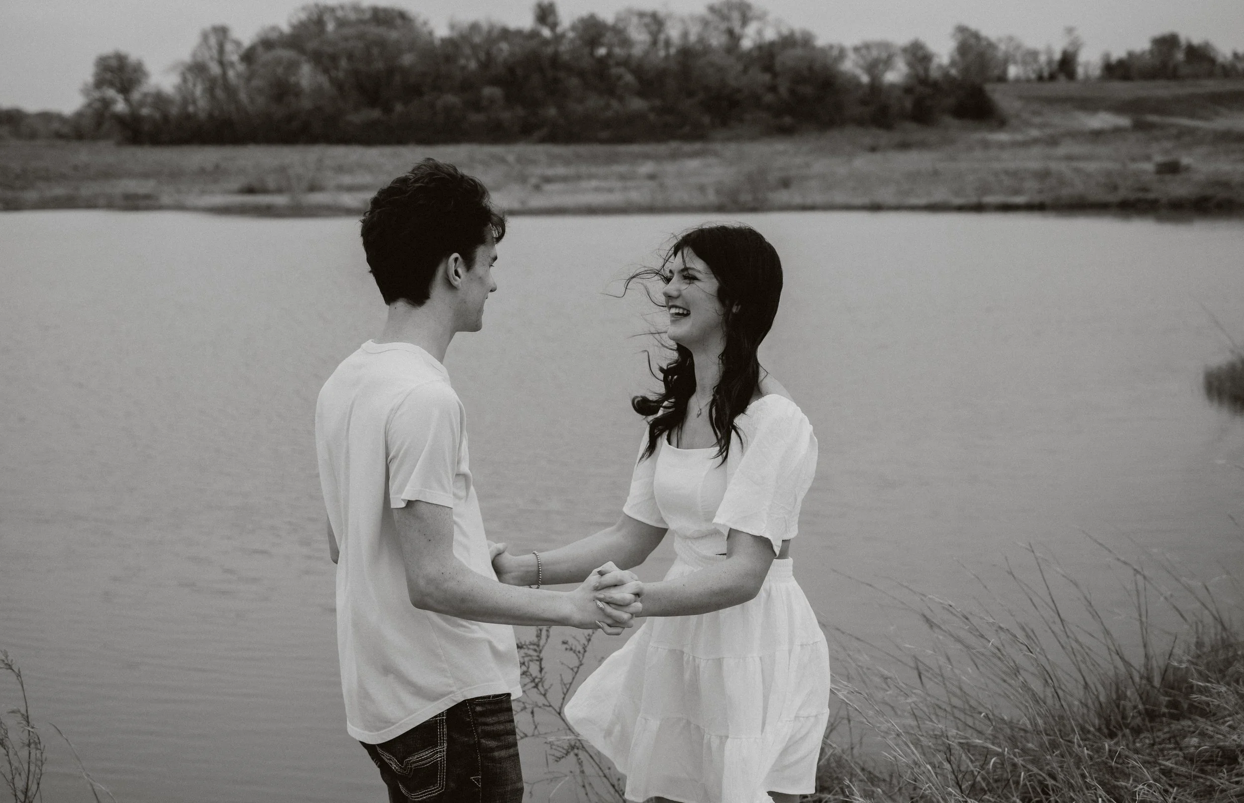 Black and white senior portrait of a couple holding hands by a lakeside, captured with intimacy and natural connection.