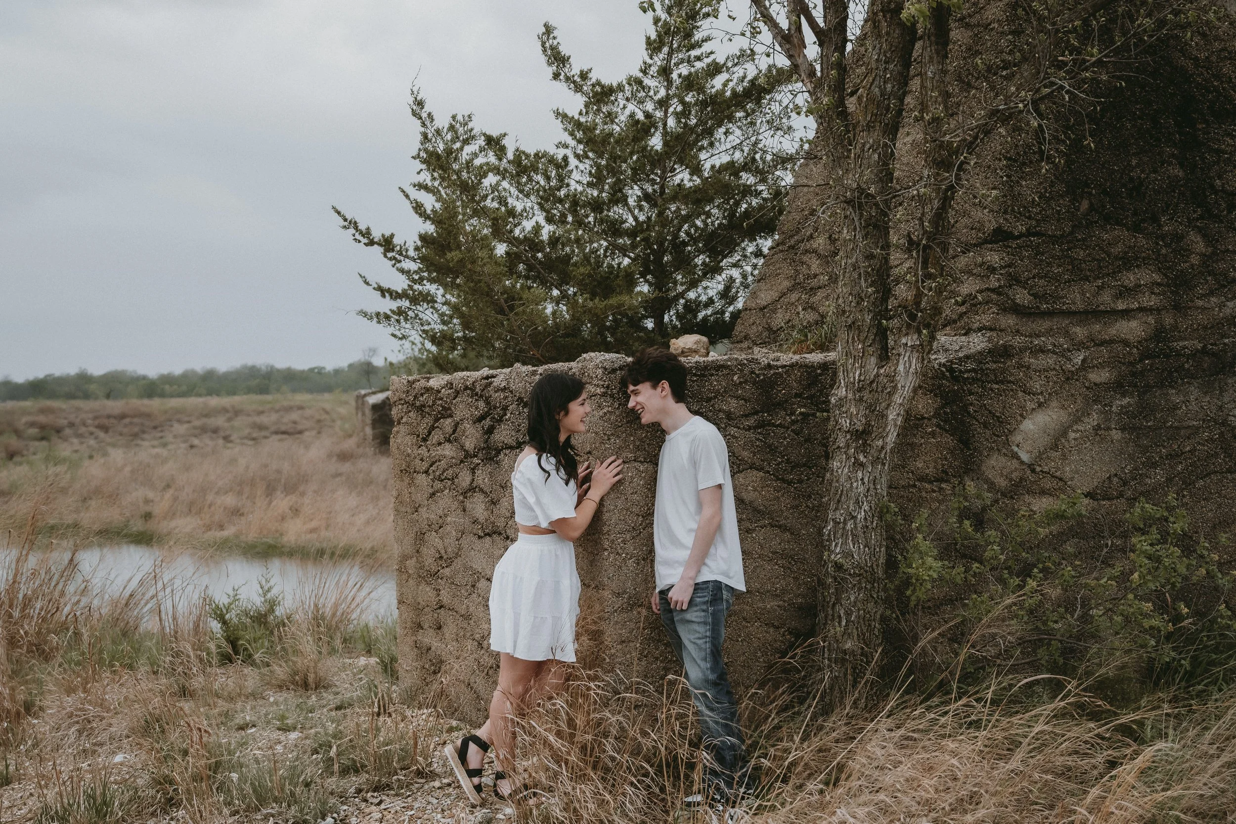A young couple standing close to each other outdoors near a rustic stone and wooden structure, smiling and engaging in conversation, surrounded by dry grass and trees.