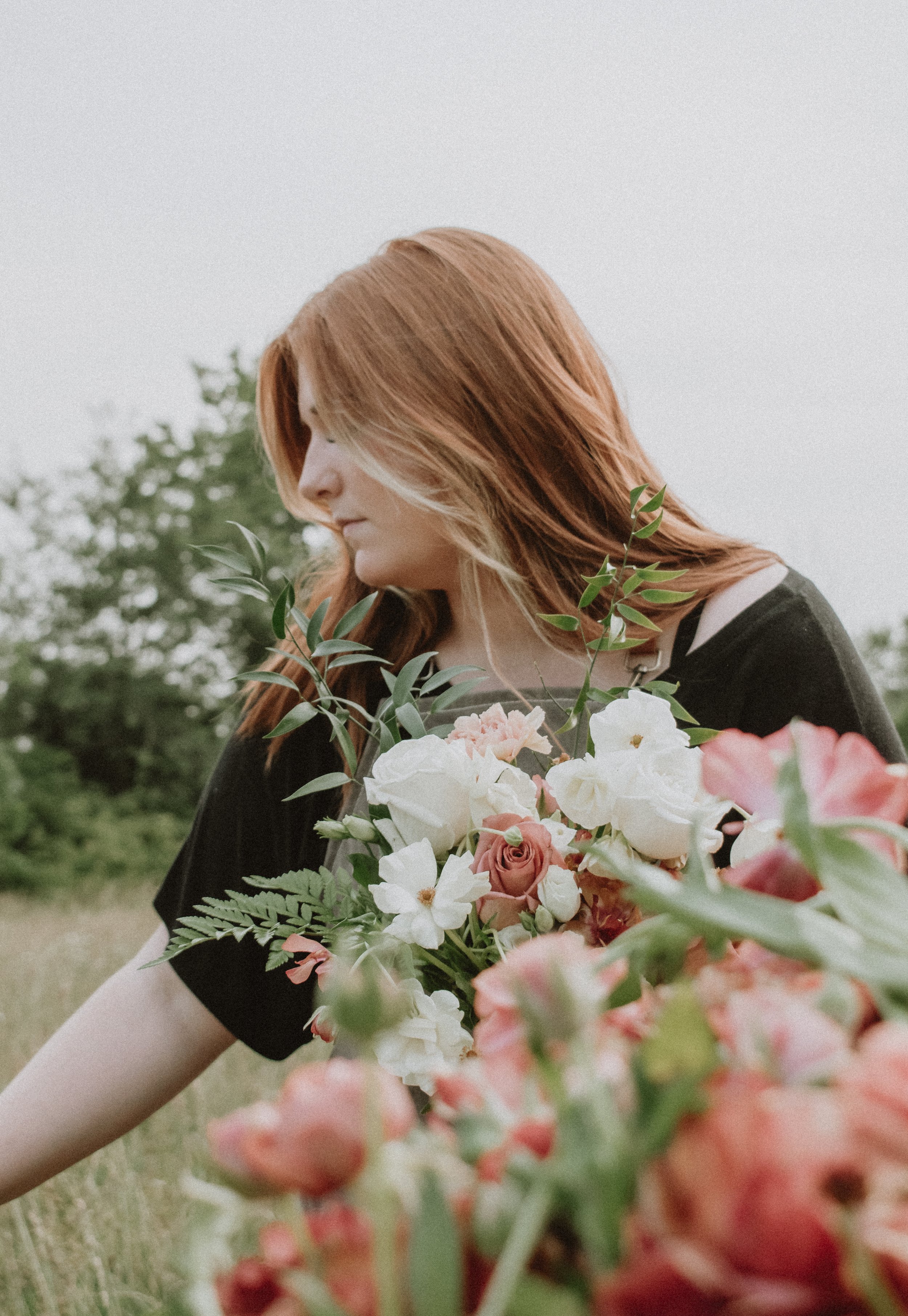 A woman with long red hair holding a bouquet of white, pink, and peach flowers in an outdoor setting.