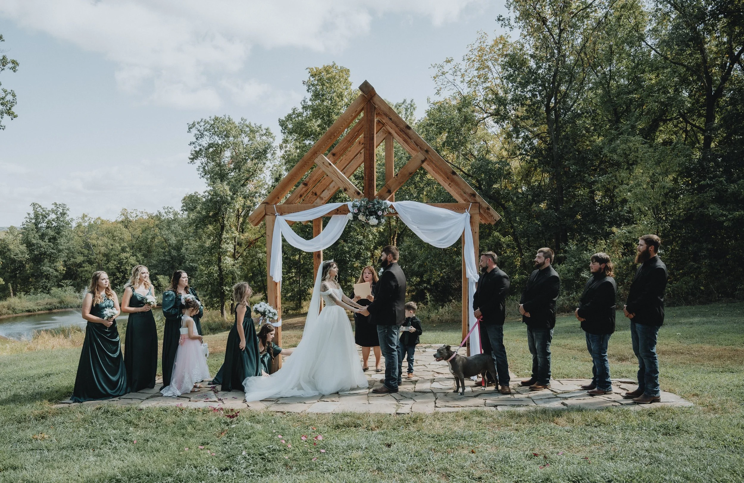 Outdoor wedding ceremony at a riverside venue in the Ozarks, bride and groom exchanging vows beneath a wooden arch.