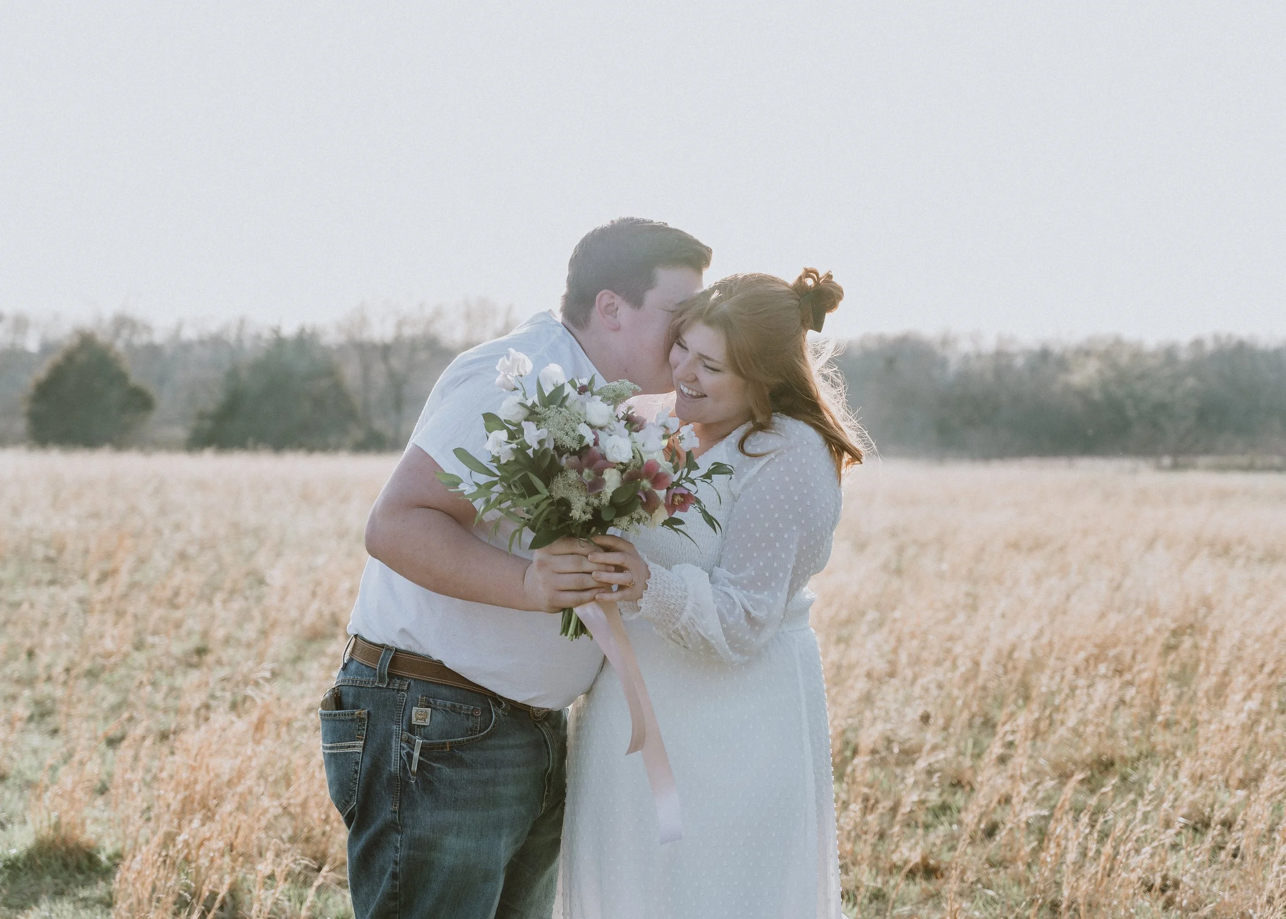 A couple standing in a field, sharing a moment, with the woman holding a bouquet of flowers and both smiling.