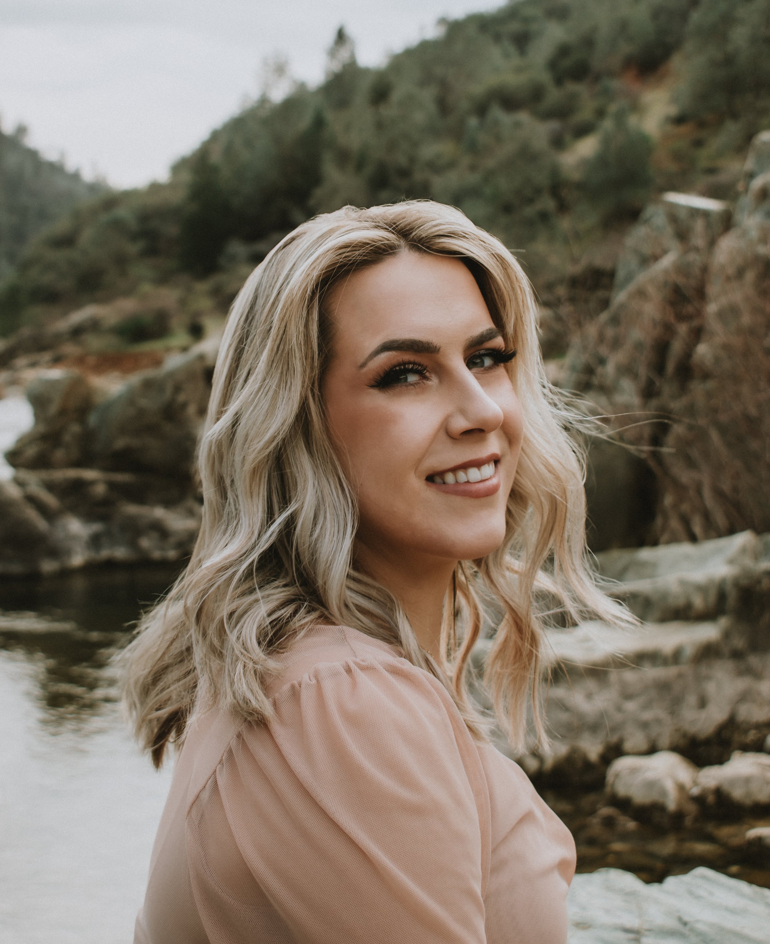 A young couple standing close to each other outdoors near a rustic stone and wooden structure, smiling and engaging in conversation, surrounded by dry grass and trees.