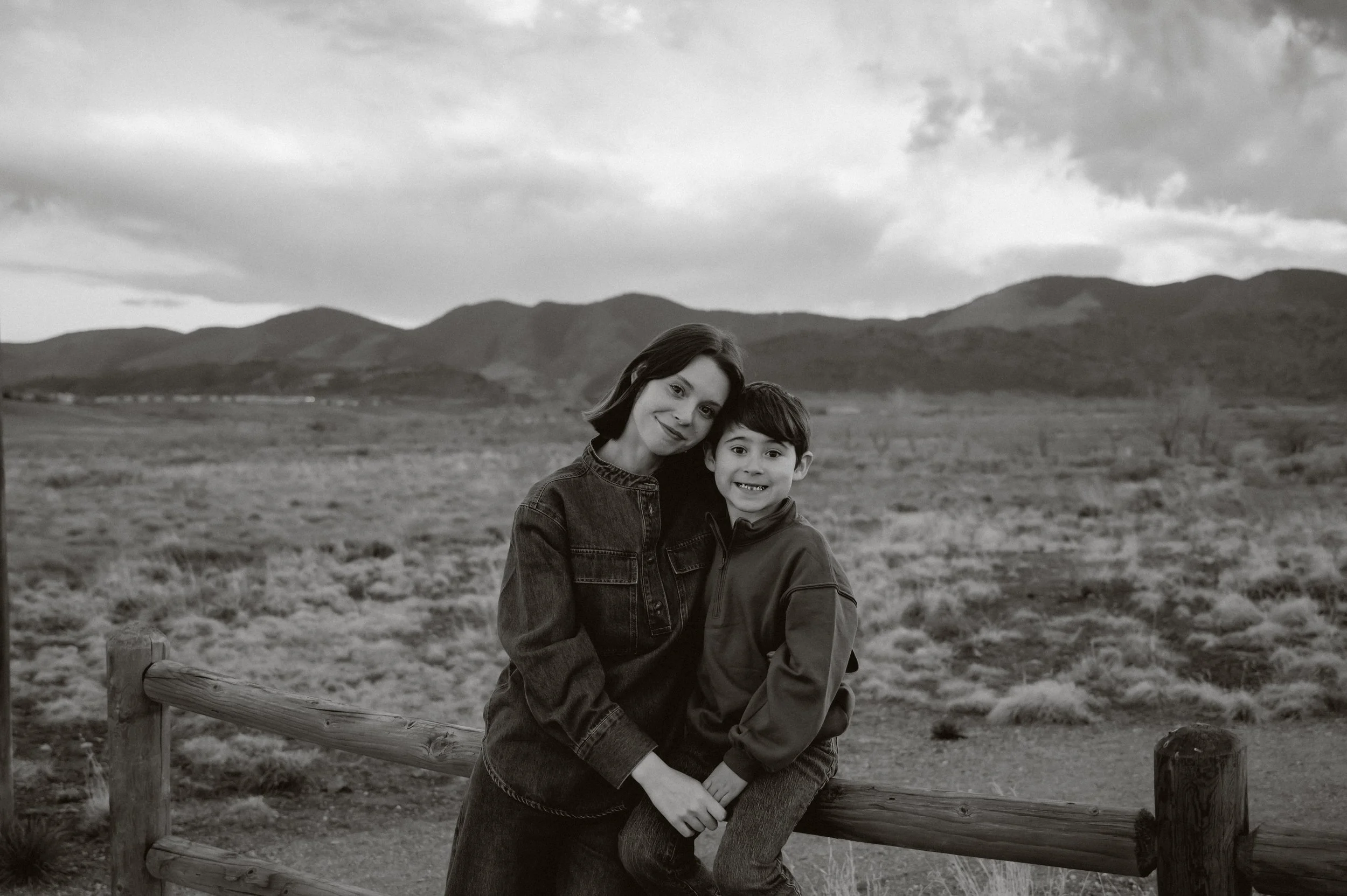 Black and white family portrait of a mother and child embracing outdoors, framed by open land and distant mountains.