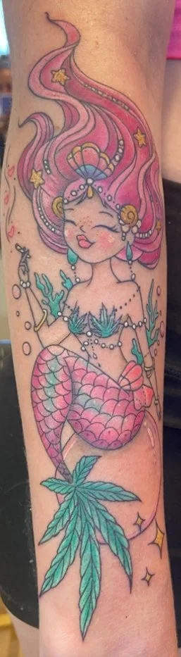 Colorful tattoo of a mermaid with pink hair, seashell bra, and a leafy tail, surrounded by stars and underwater elements.