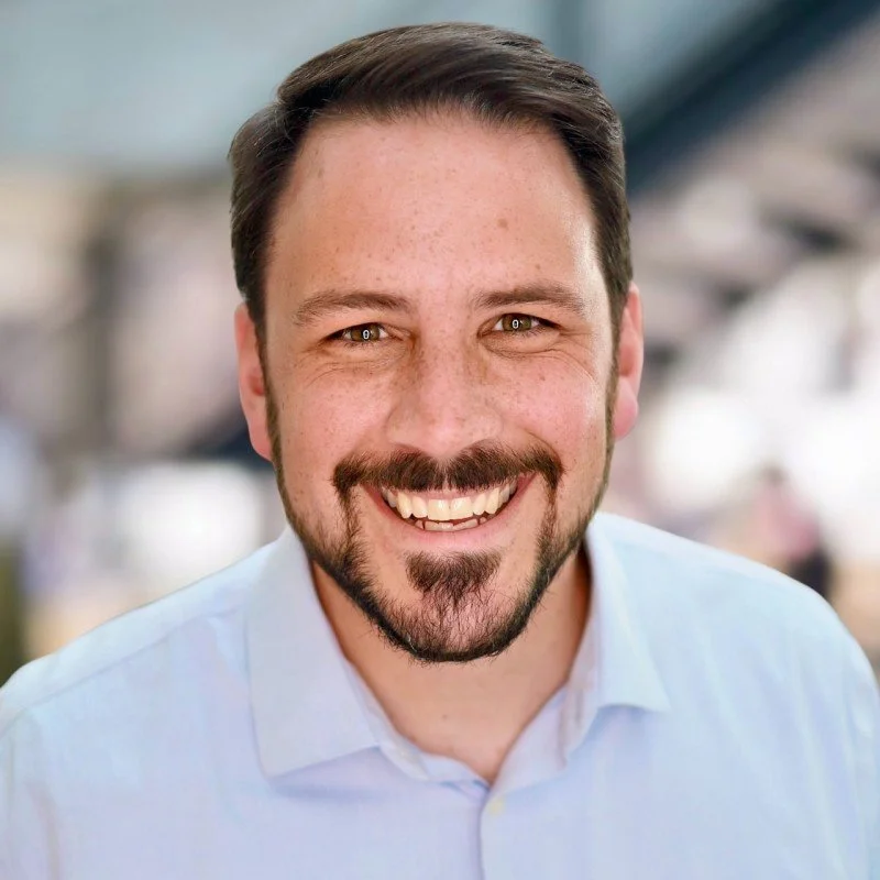 A smiling man with dark hair, beard, and mustache wearing a light blue shirt.