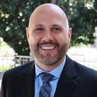 A smiling man with a beard and bald head wearing a navy blazer, light blue shirt, and patterned tie, outdoors with greenery in the background.