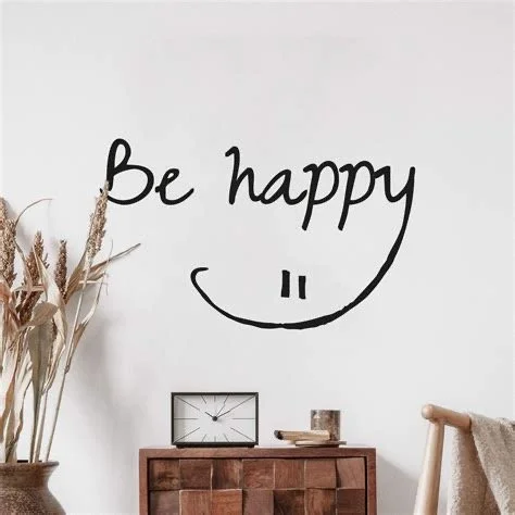 White wall with black handwritten message 'Be happy' and a smiley face. There is a wooden table with a clock, book, and small cup, and a dried plant in a vase.