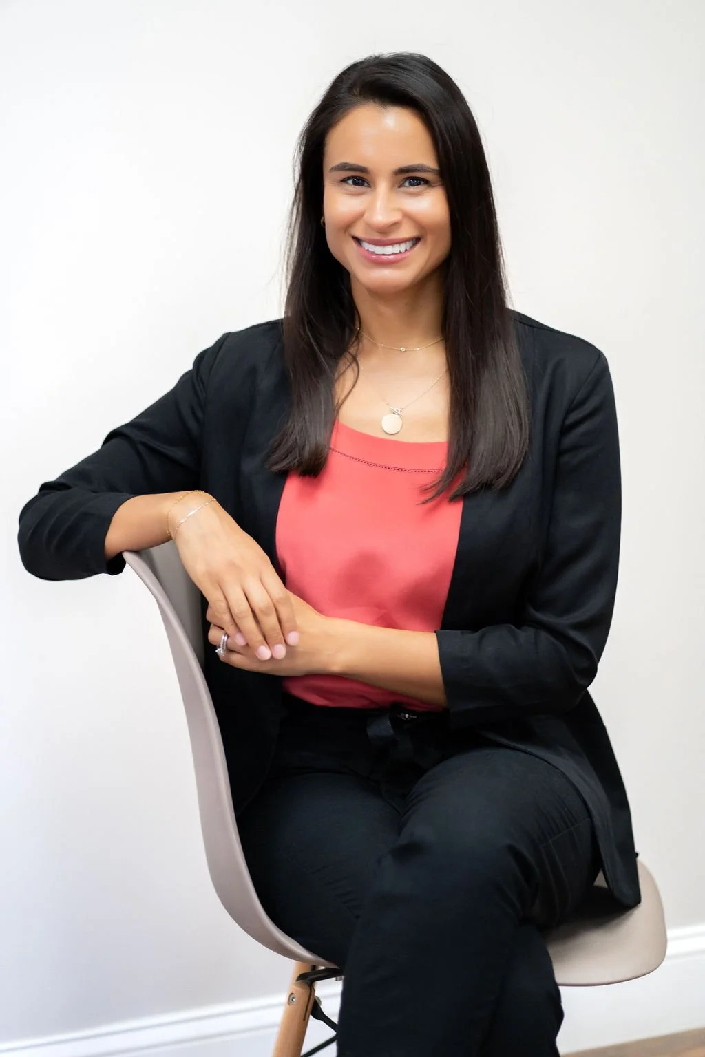 A young woman with long dark hair, sitting on a chair, smiling, wearing a black blazer over a coral top, with jewelry, against a white background.