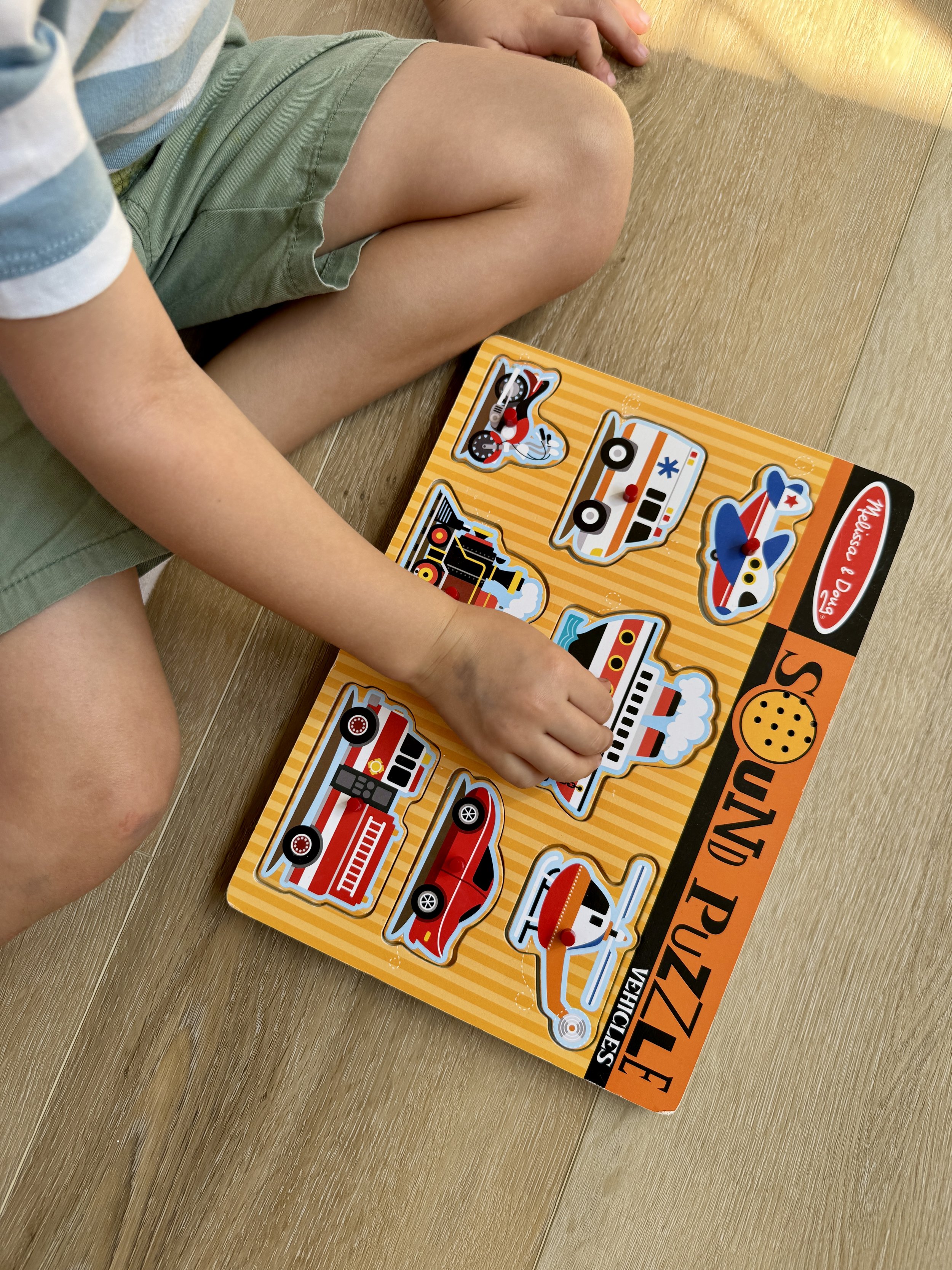 Child playing with a vehicle-themed puzzle on a wooden floor, with various vehicle pieces such as a fire truck, race car, and airplane.