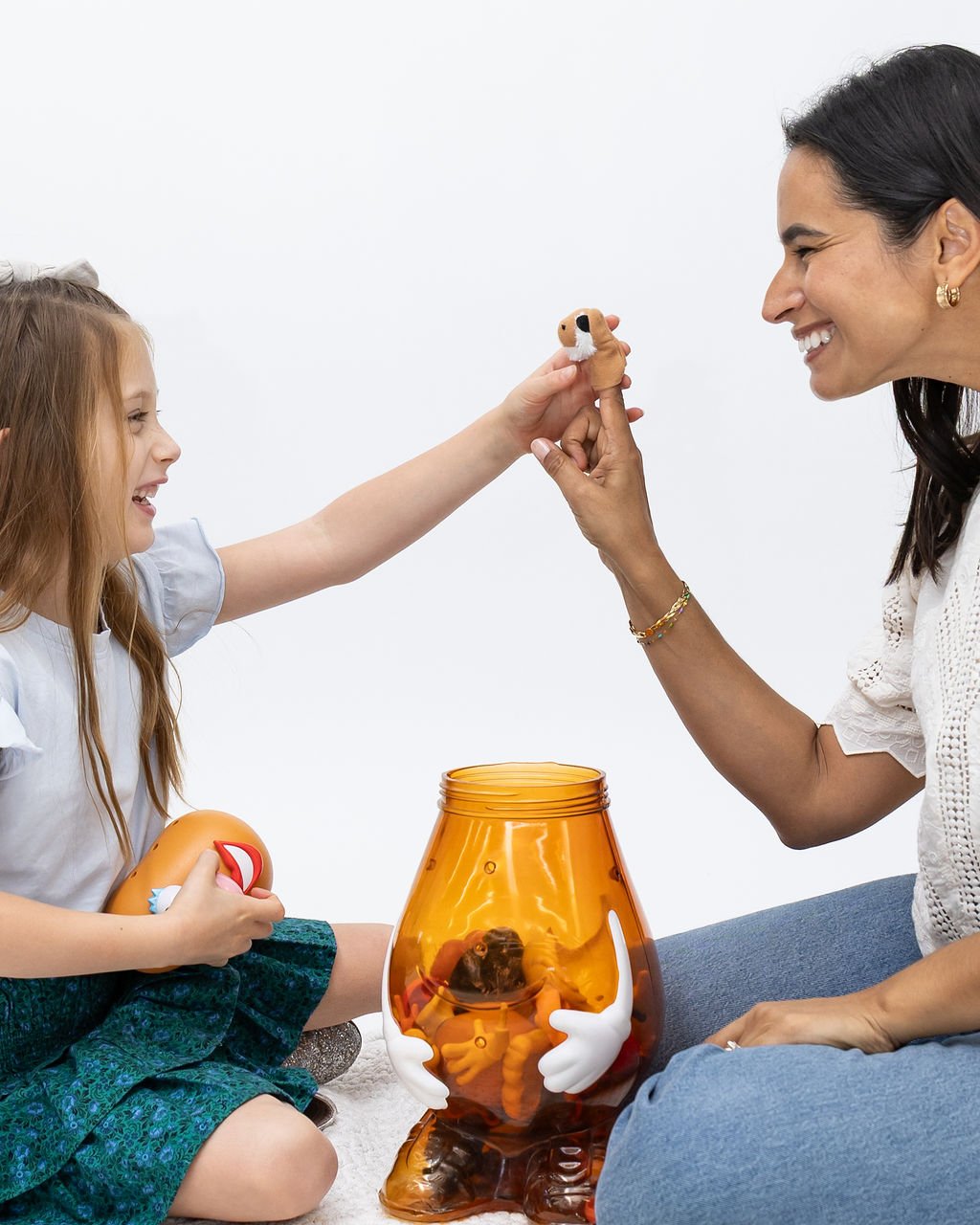 A woman and a girl playing with a small dog plush toy. The woman is holding the plush dog close to the girl's face, and they are smiling at each other. The girl is holding a stuffed animal with a balloon in her lap. There is an amber-colored fish bowl with a white glove-shaped hand inside it, resembling a character or decoration.