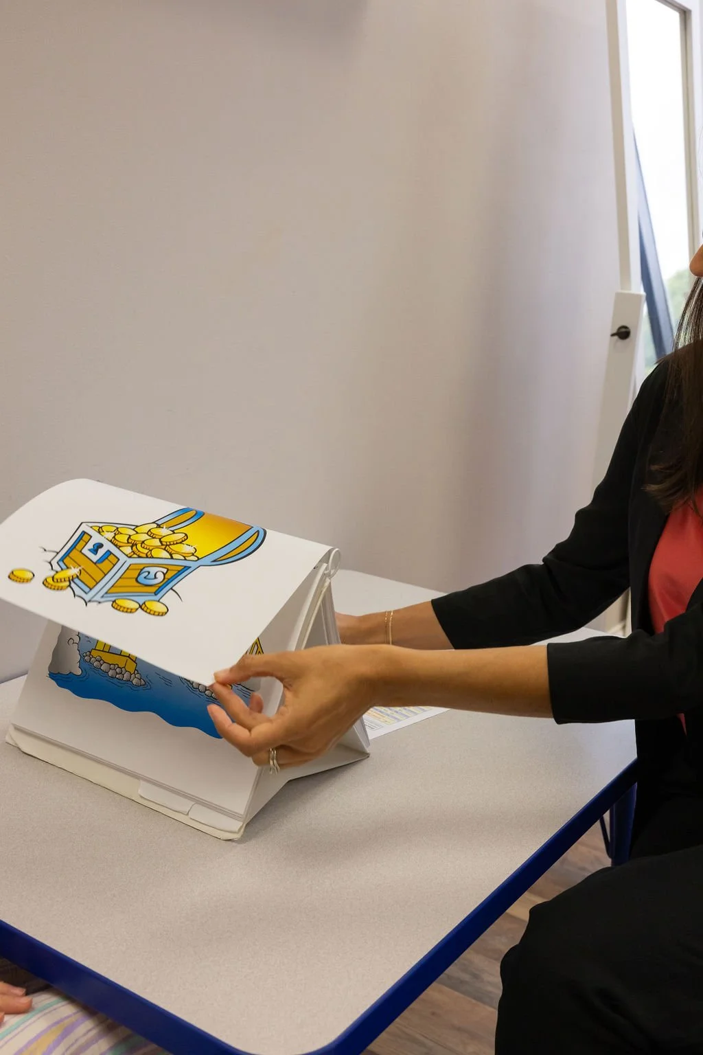 A woman holding an illustrated book with images of a treasure chest and a ship in a classroom or office setting.