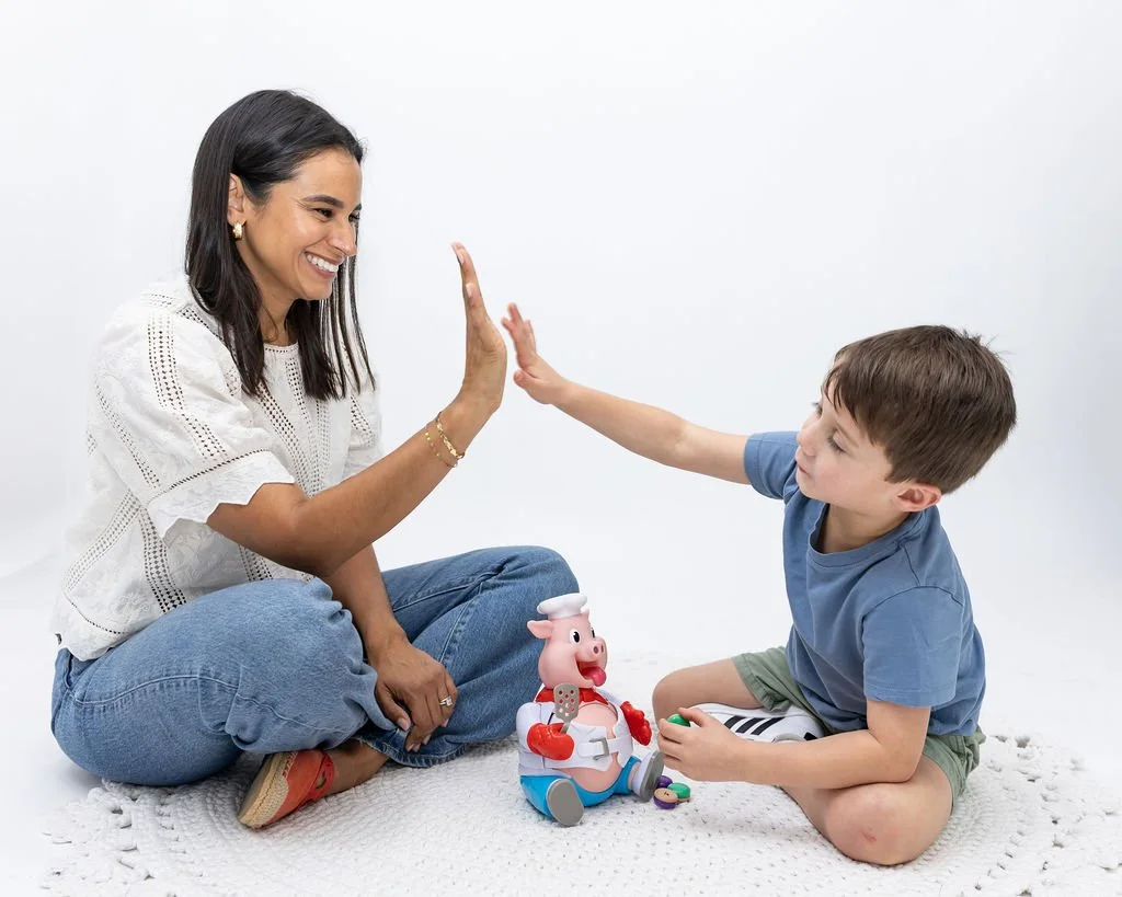 A woman and a young boy sitting on a white blanket, giving a high five together, with a toy pig dressed as a chef between them.