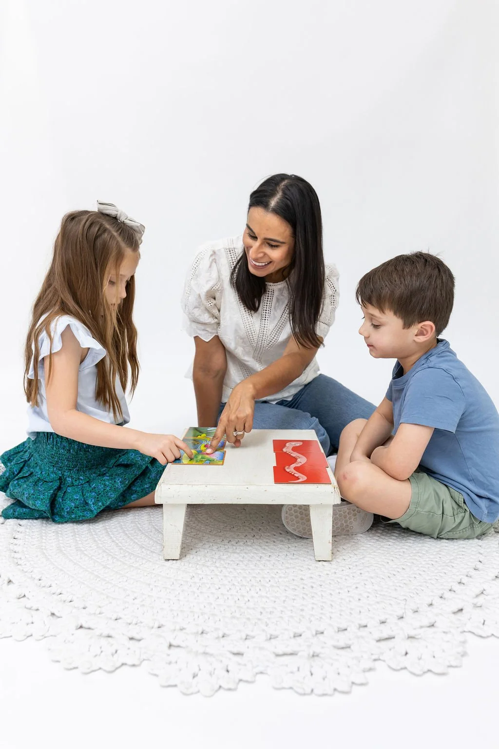 Woman playing a board game with two children on a small white table in a minimalistic room.