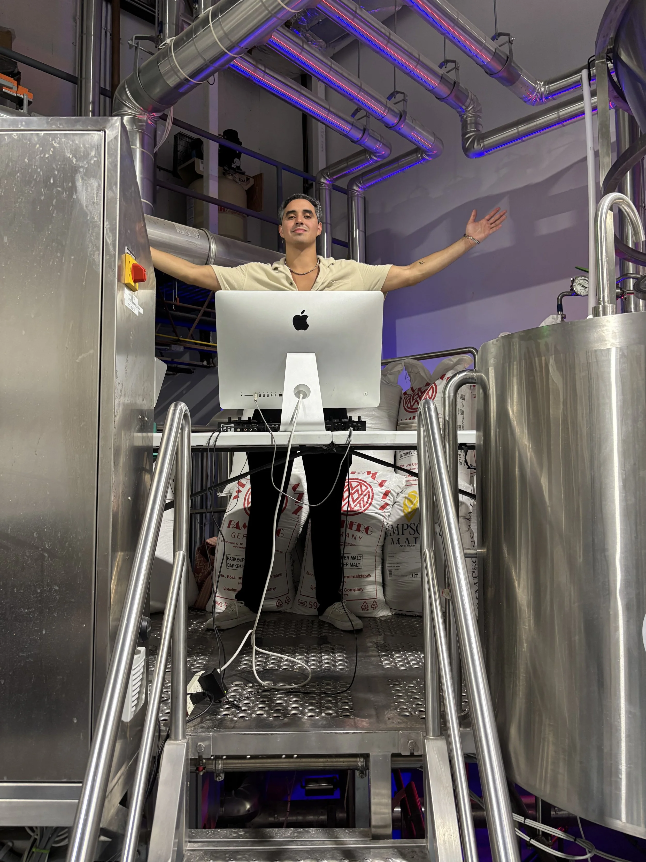 A DJ stands on a metal platform in an industrial setting with pipes overhead, using a computer with arms stretched out in a celebratory gesture.