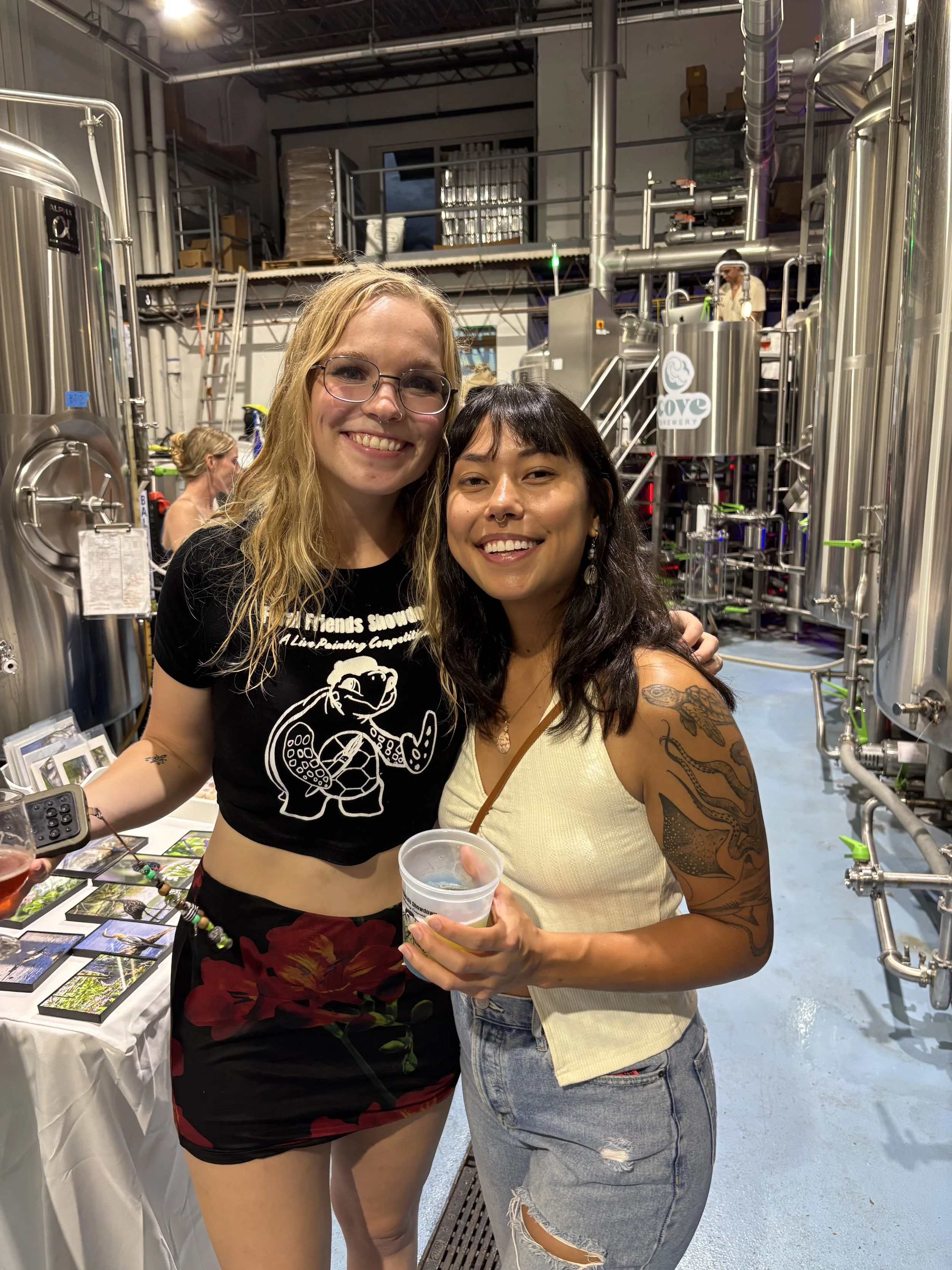 Two women smiling and posing together inside a brewery, with large stainless steel tanks and brewing equipment in the background. One woman has blonde hair, wearing glasses, and a black t-shirt with a graphic. The other woman has dark hair, visible t