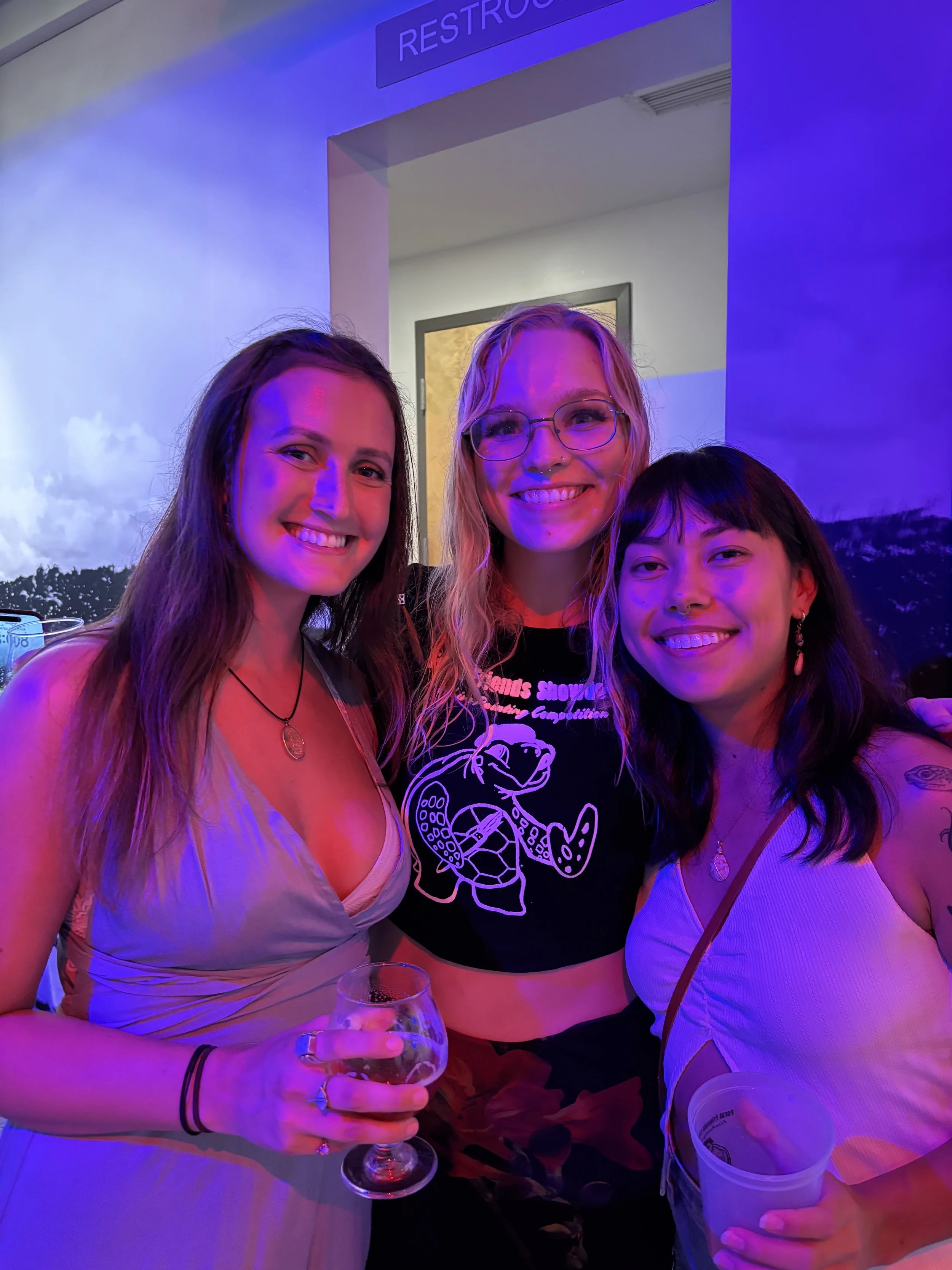 Three smiling women at a party standing closely together under purple lighting, with drinks in their hands and a
