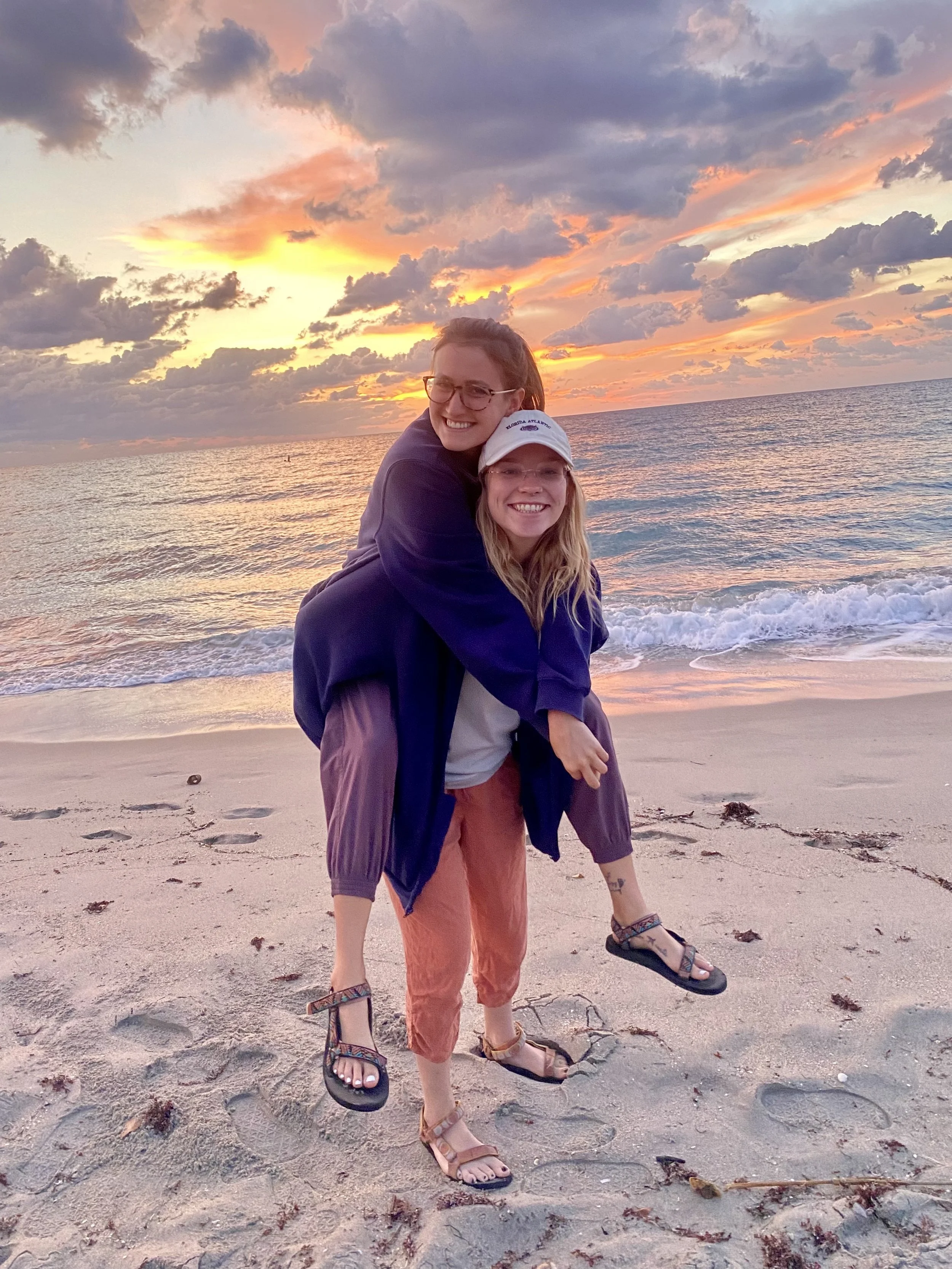 Two women smiling and hugging on a beach at sunset, with clouds and the ocean in the background.