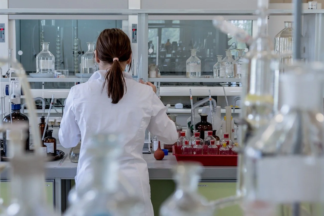 A scientist with brown hair tied in a ponytail, wearing a white lab coat, working with glass bottles and laboratory equipment in a laboratory.
