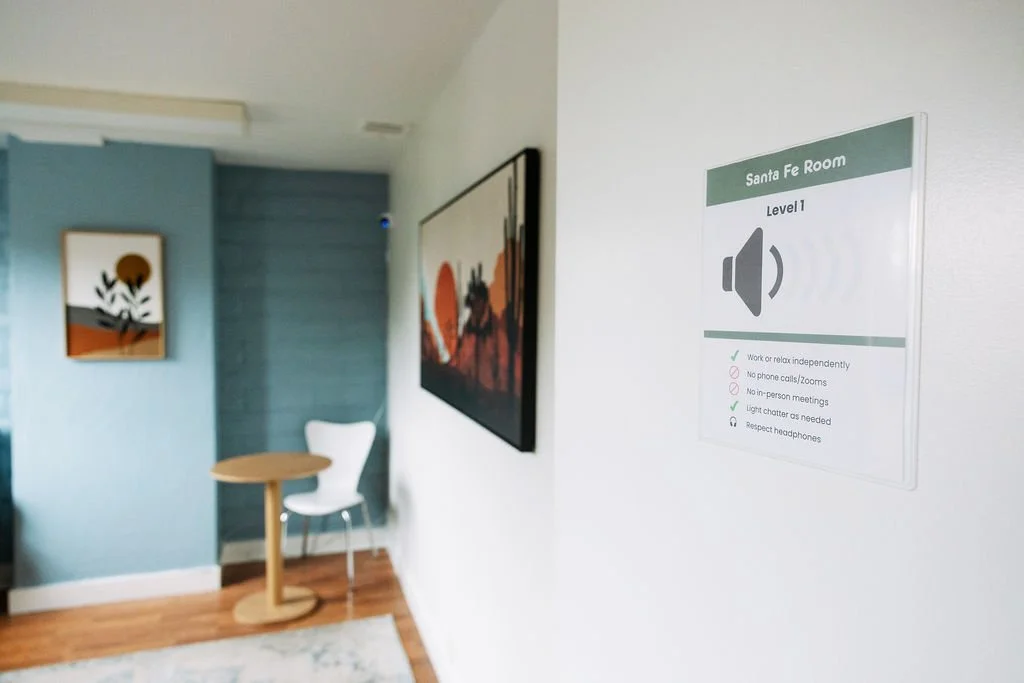A white wall with a sign for the Santa Fe Room, level 1, indicating rules like no phone calls or in-person meetings, and respecting headphones. Part of an interior space with wall art, a small table, and a white chair.