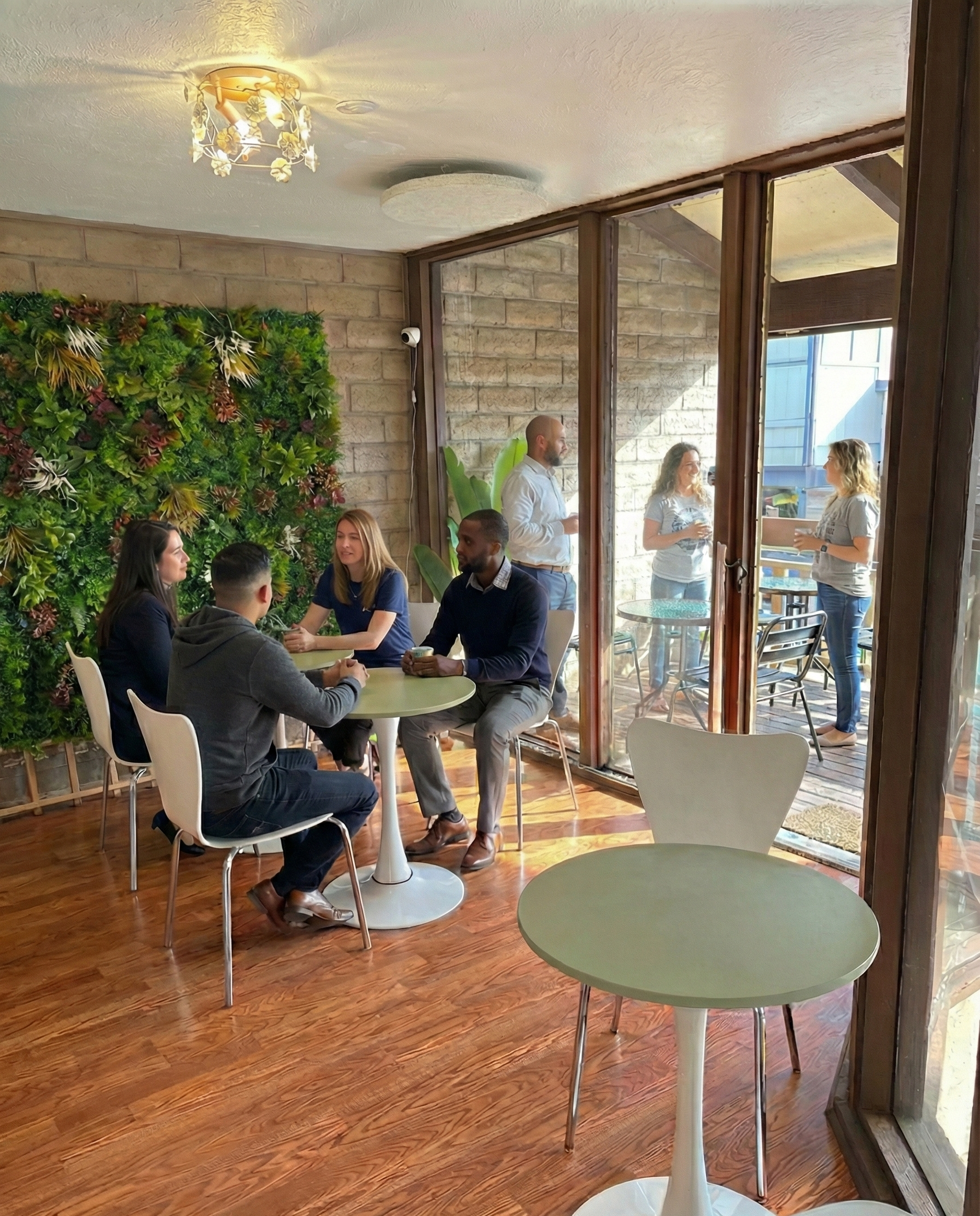 A group of five people sitting around a table inside a cafe, with two women and three men, some holding drinks and one using a phone. Outside through the large glass doors, three women are standing and talking on a patio with outdoor furniture.