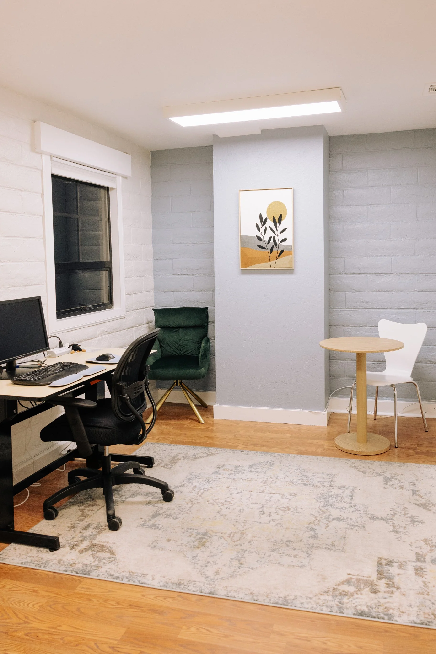 Office space with a desk and computer, green armchair, white and wooden table, textured gray and white walls, a framed art piece, window, and ceiling light.