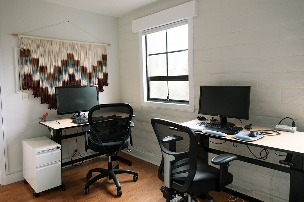 Two office desks with monitors, keyboards, and office chairs in a room with white brick walls, a window, and a woven wall hanging.