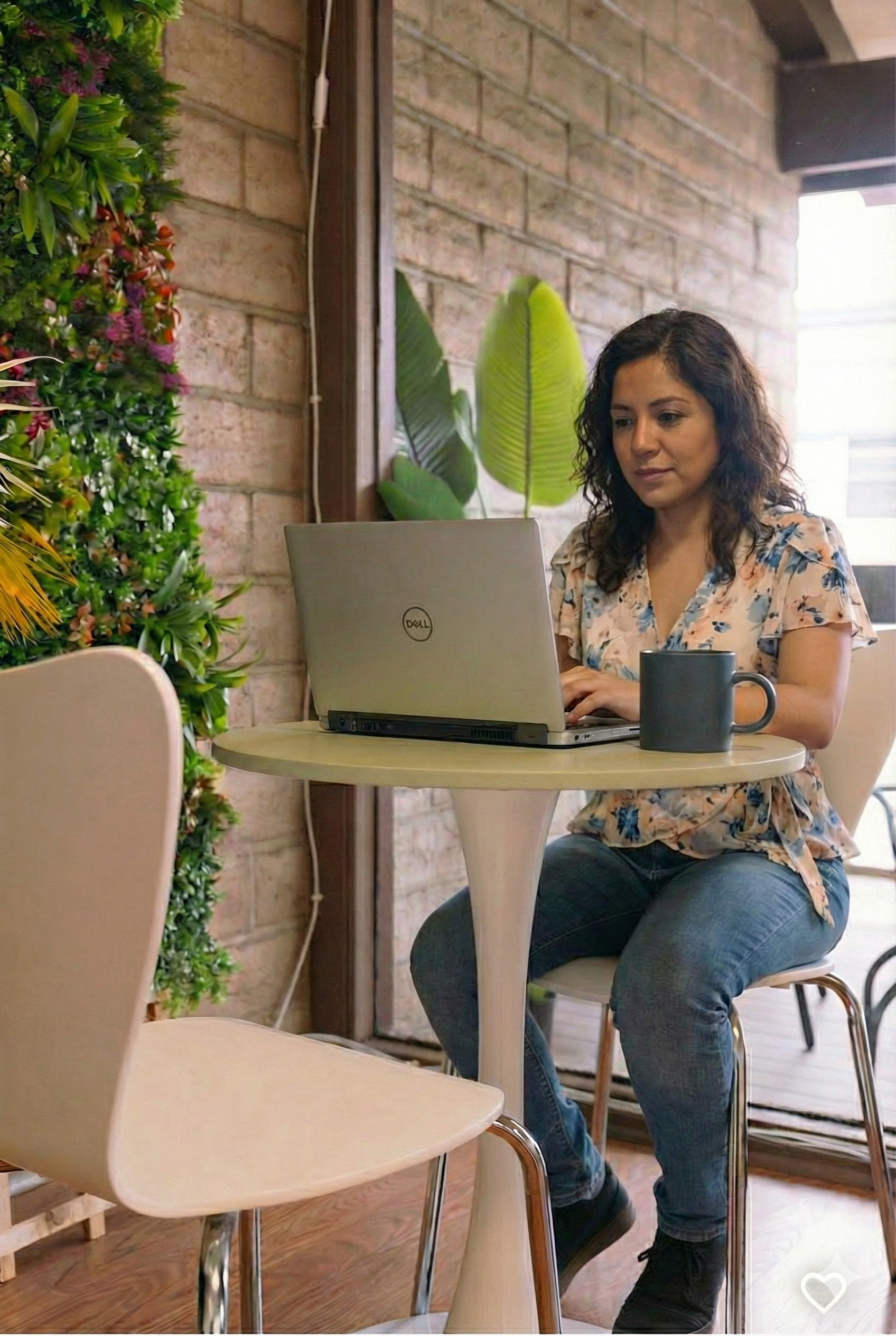 A woman working on a laptop at a small round table in a cozy cafe with a brick wall and green plants in the background.