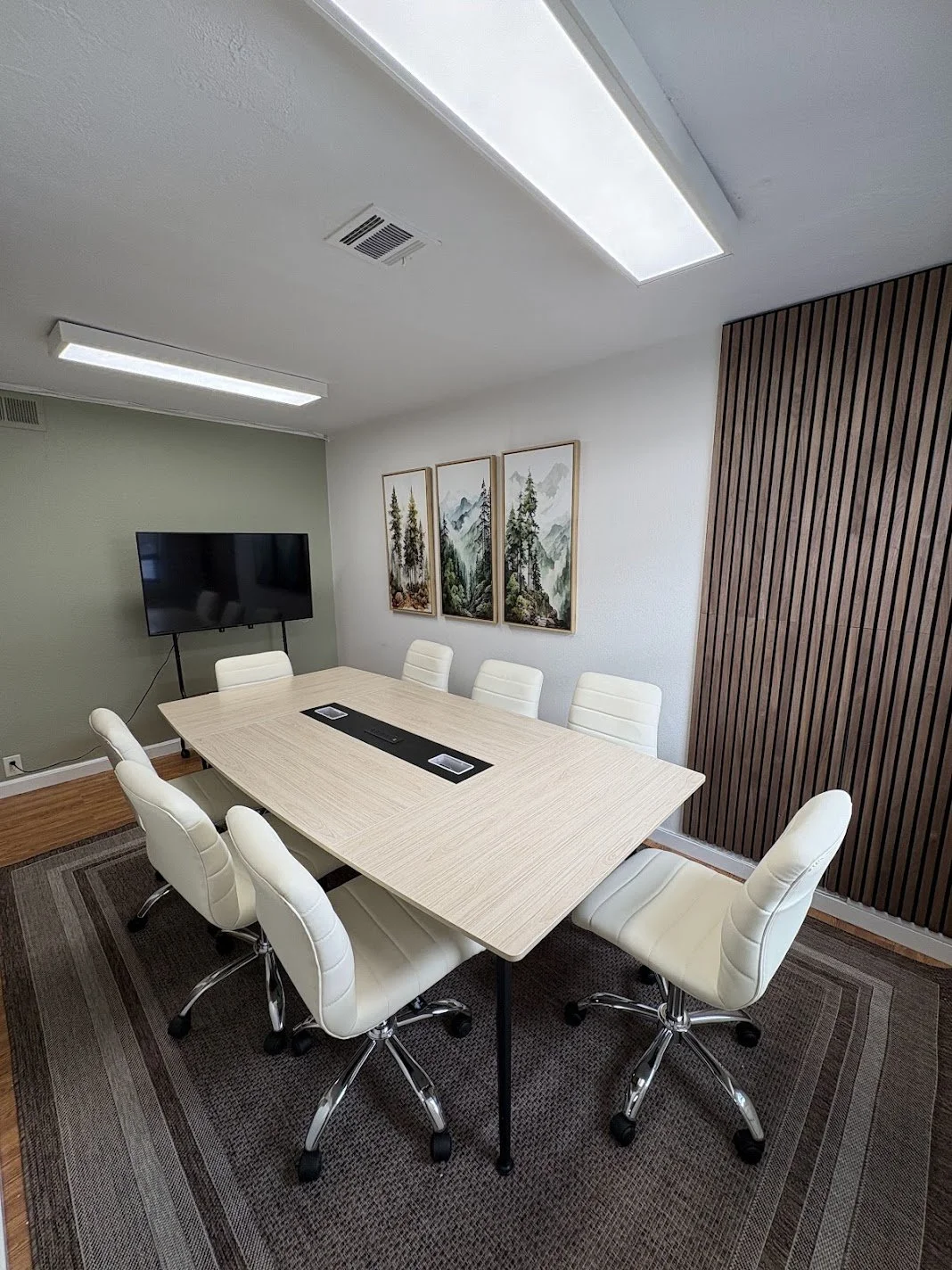 A modern conference room with a light wood table, eight white chairs, a wall-mounted flat-screen TV, three framed landscape paintings, a dark wood slatted accent wall, and ceiling lights.