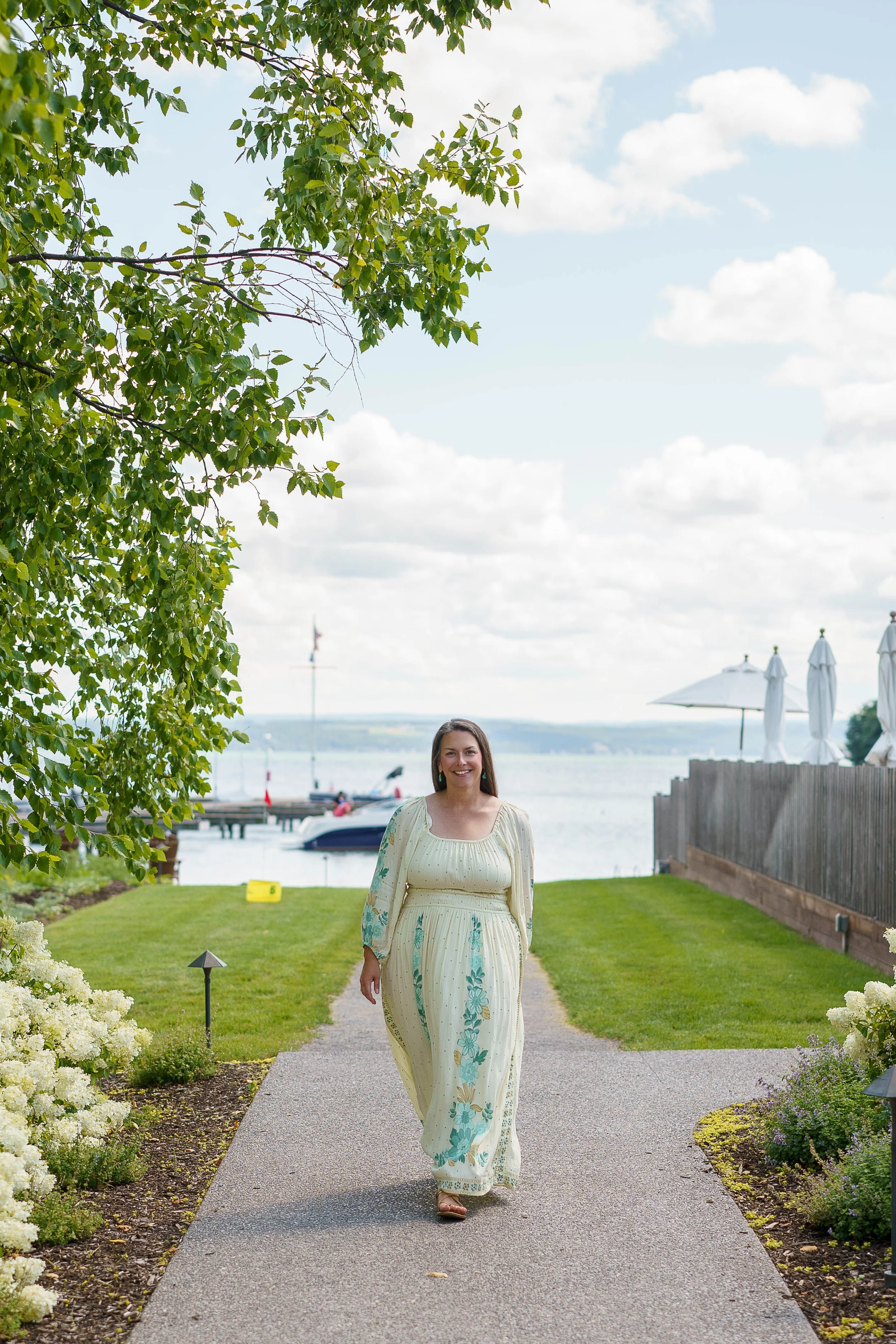 Aubree Guiffre, perimenopause and menopause coach, walking on a garden path near a waterfront with boats and umbrellas, smiling, dressed in a long cream-colored dress with teal floral patterns, surrounded by white flowers and green trees.